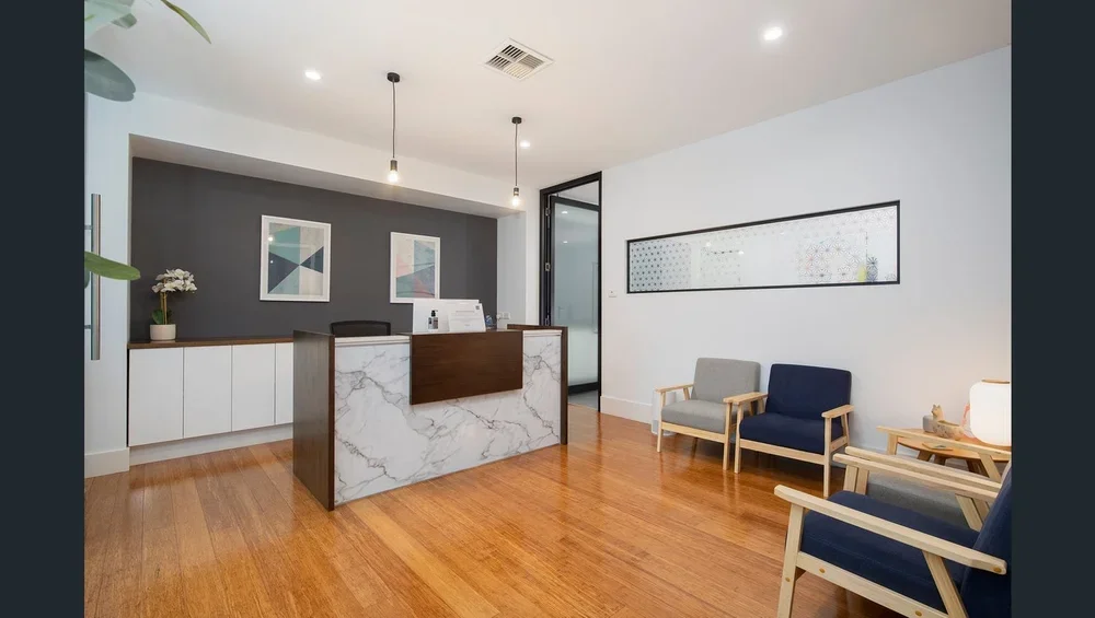 Interior of a modern reception area with a wooden floor, white and gray walls, a reception desk with a marble top, and three chairs with wooden frames and cushions, along with a side table with a lamp.