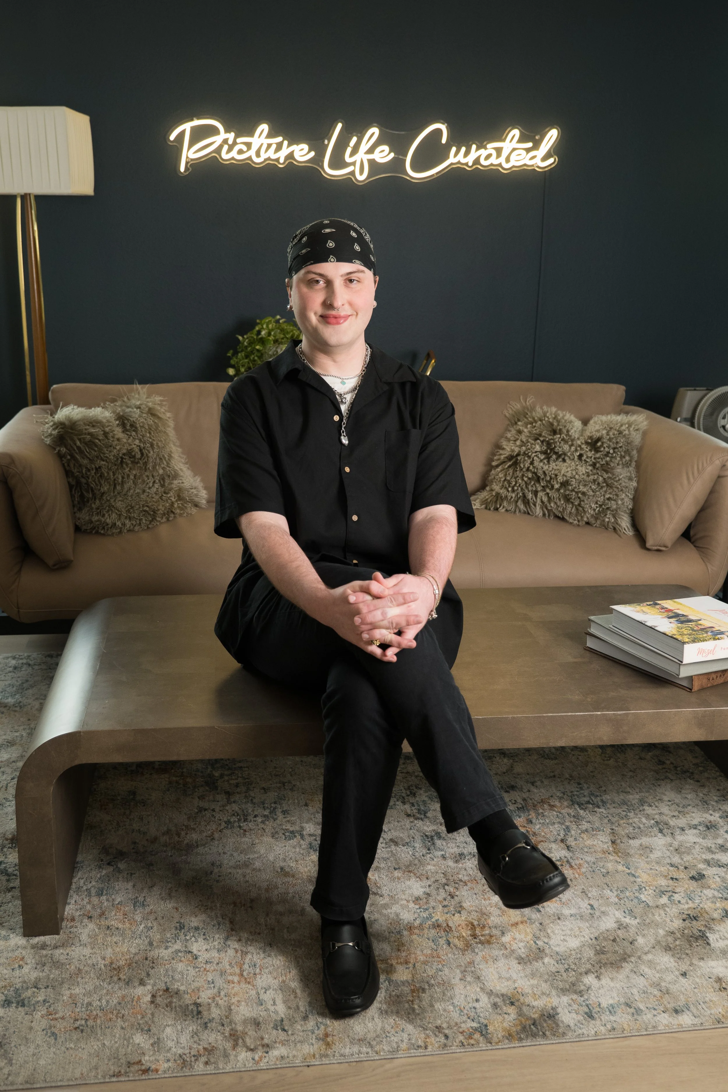 Person sits on a coffee table in a modern living room with a navy painted wall that features a neon sign reading 'Picture Life Curated'. The person wears a black bandana, black shirt, and black shoes, with blonde hair and jewelry. A beige sofa with fur pillows and a stack of magazines are visible.