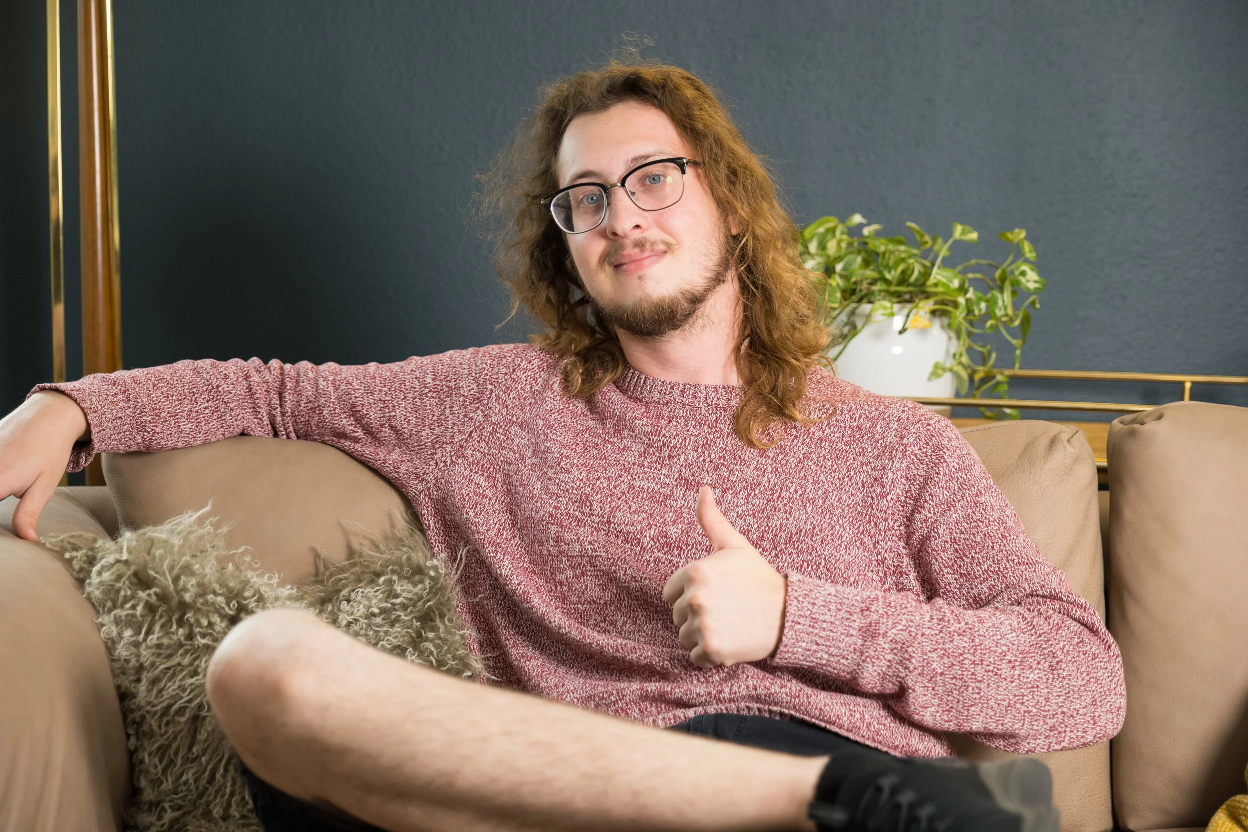 A young man with long curly hair, glasses, and a beard sitting on a beige couch, giving a thumbs-up gesture.