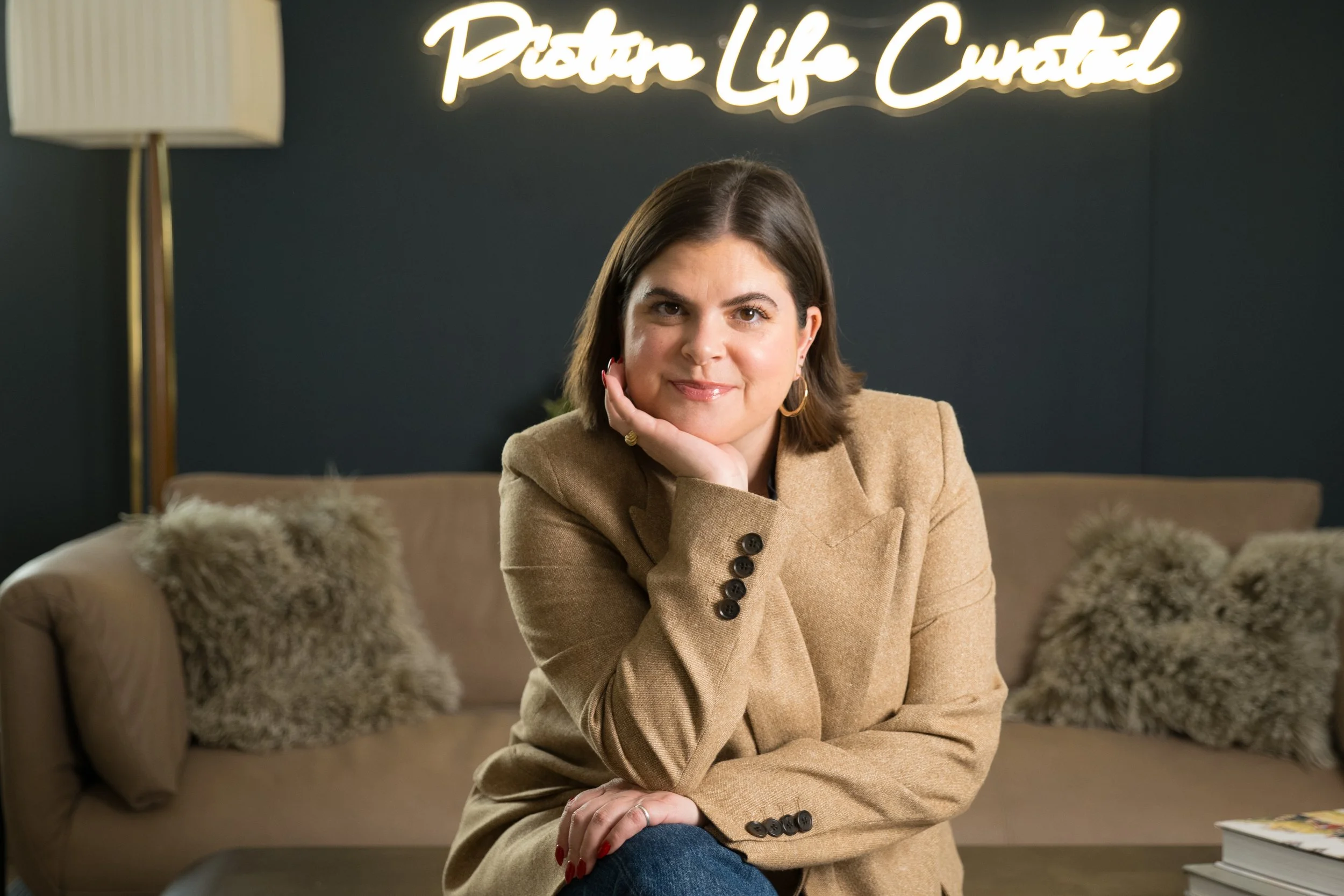 A woman with short brown hair and gold hoop earrings sitting on a beige sofa, resting her chin on her hand, in a cozy modern room with a dark wall, fluffy pillows, a tall floor lamp, and a neon sign that reads 'Picture Life Curated'.