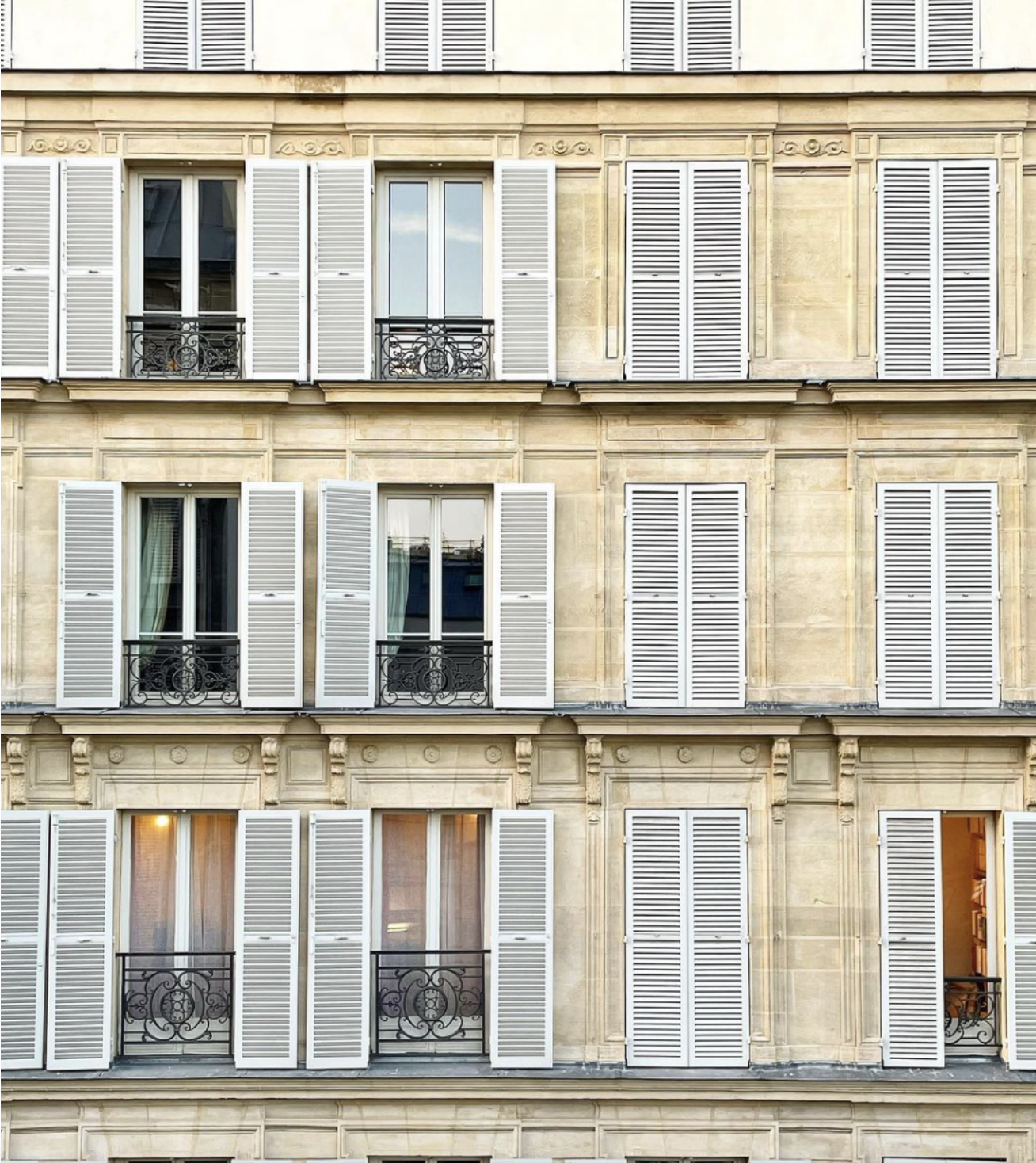 Facade of a Parisian building with four floors, each featuring a row of windows with white shutters and small balconies with ornate black iron railings.
