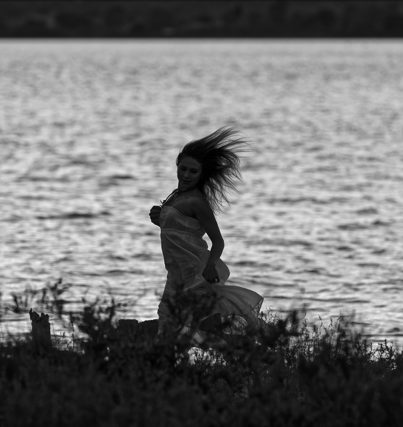A black-and-white photo of a woman in a flowing dress standing by water with her hair blowing in the wind.