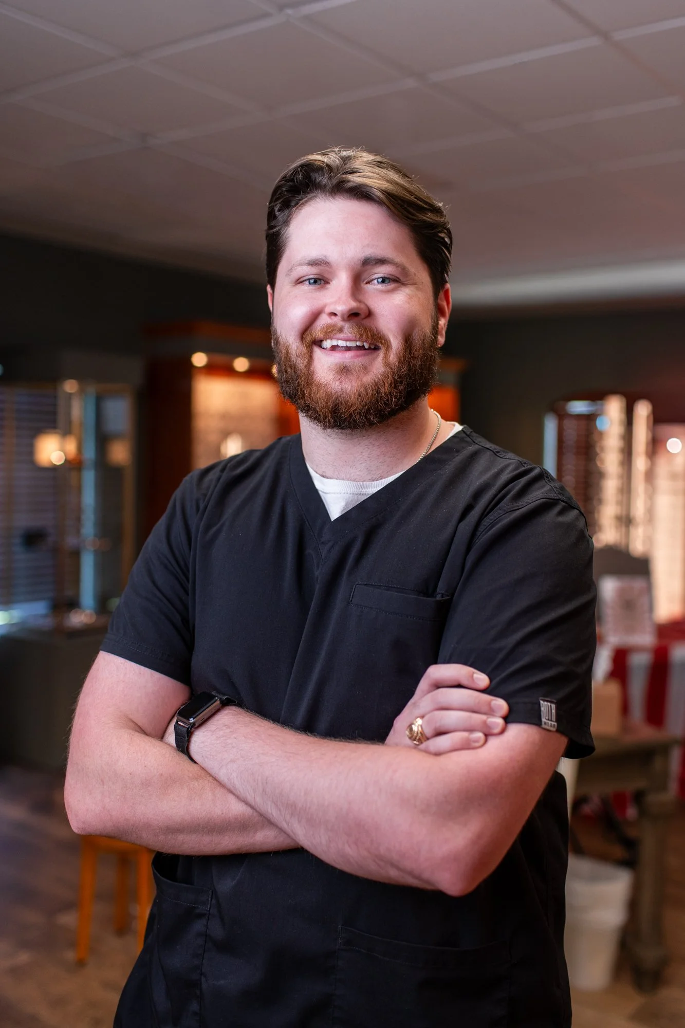 Dr Jadon Borders. An eye doctor in Longview Tx. A smiling man with a beard and short brown hair, wearing a black shirt, standing with crossed arms in a warmly lit indoor setting.