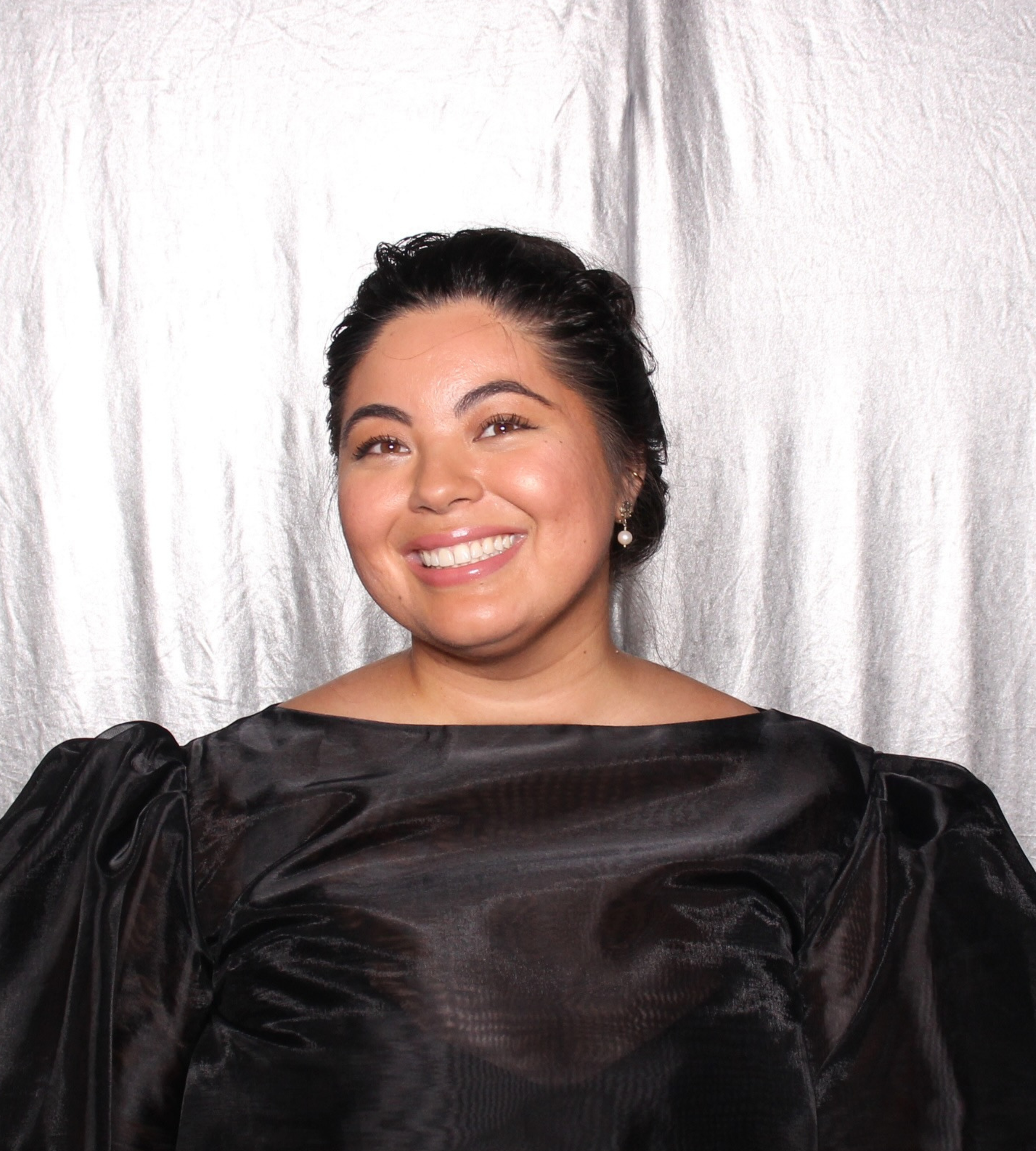 A woman smiling in front of a silver backdrop, wearing a black dress with puffed sleeves and pearl earrings.