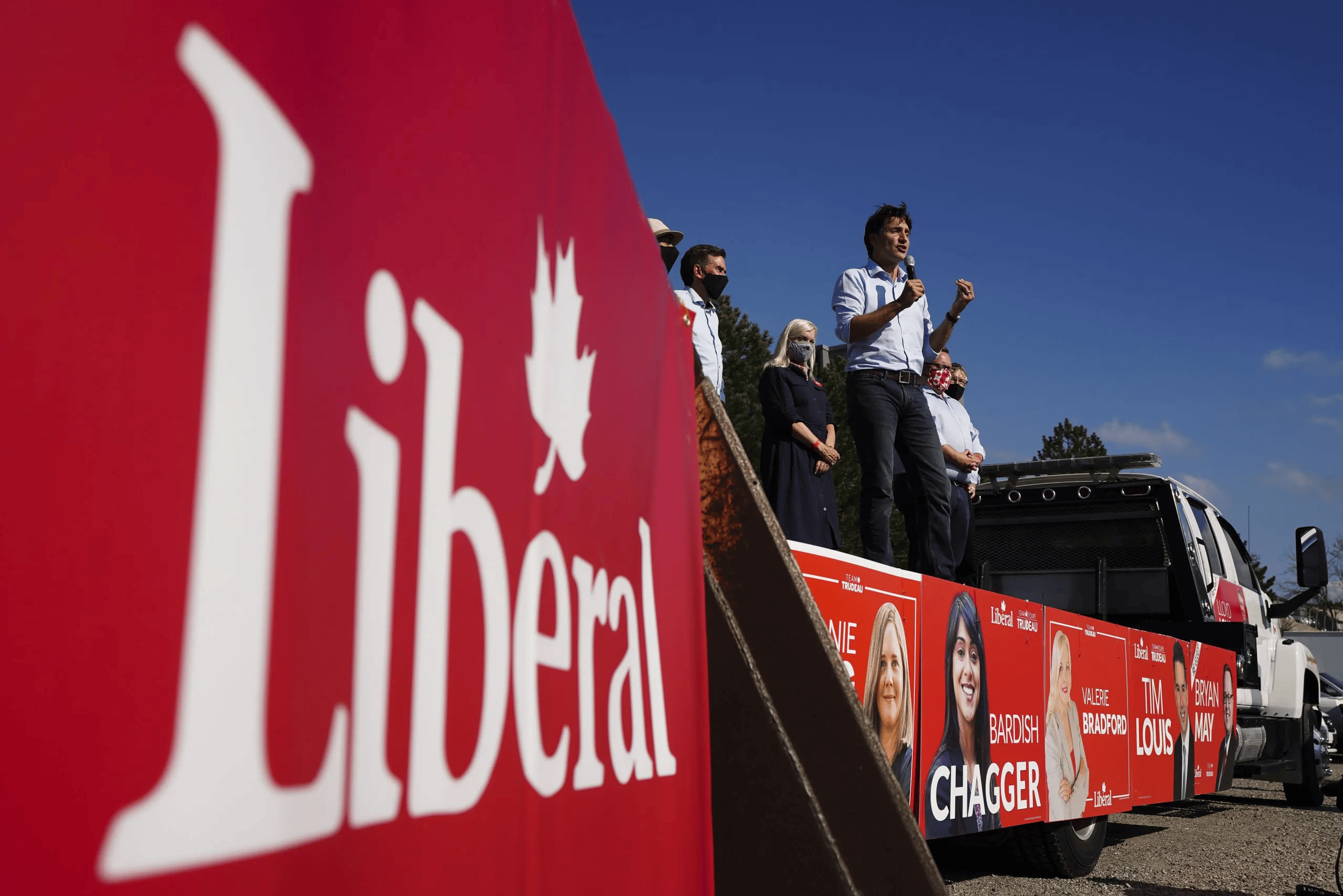 A political rally with Justin Trudeau speaking into a microphone on a stage decorated with campaign posters, and a large red banner with the word 'Liberal' in focus in the foreground.