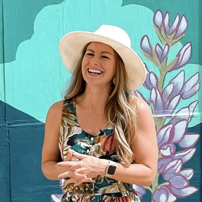 A woman with long blonde hair smiling, wearing a wide-brimmed hat and a floral dress, standing in front of a colorful mural with purple and white flowers.