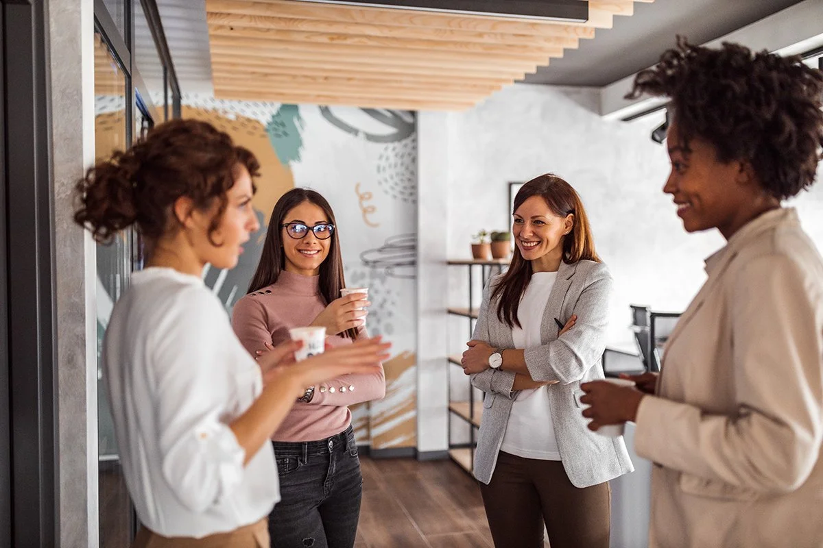 Four women having a conversation in an office or cafe, one woman is holding a cup, and they are smiling and engaged with each other.