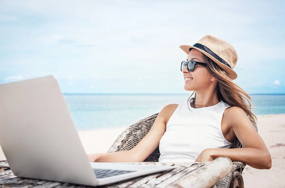 Woman wearing a straw hat and sunglasses working on a laptop while relaxing on a beach