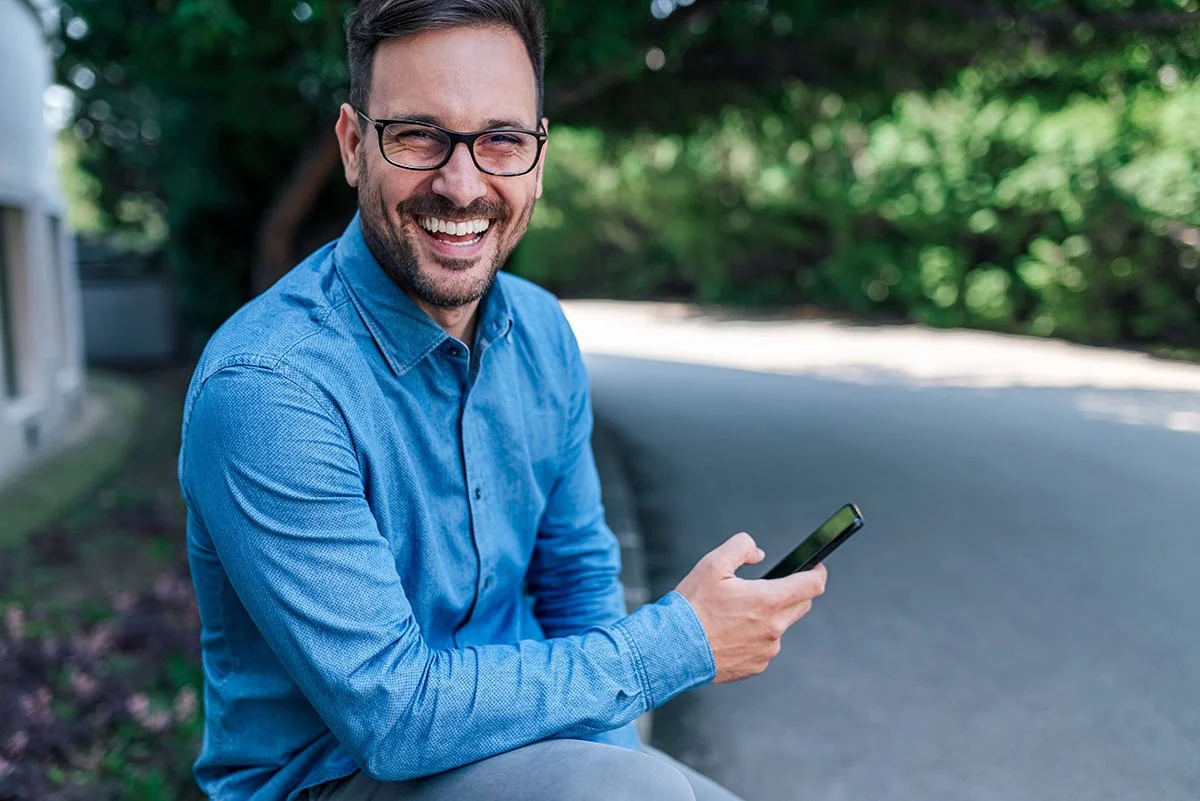 A man wearing glasses and a blue button-up shirt, smiling while looking at his phone outdoors near a sidewalk and green foliage.