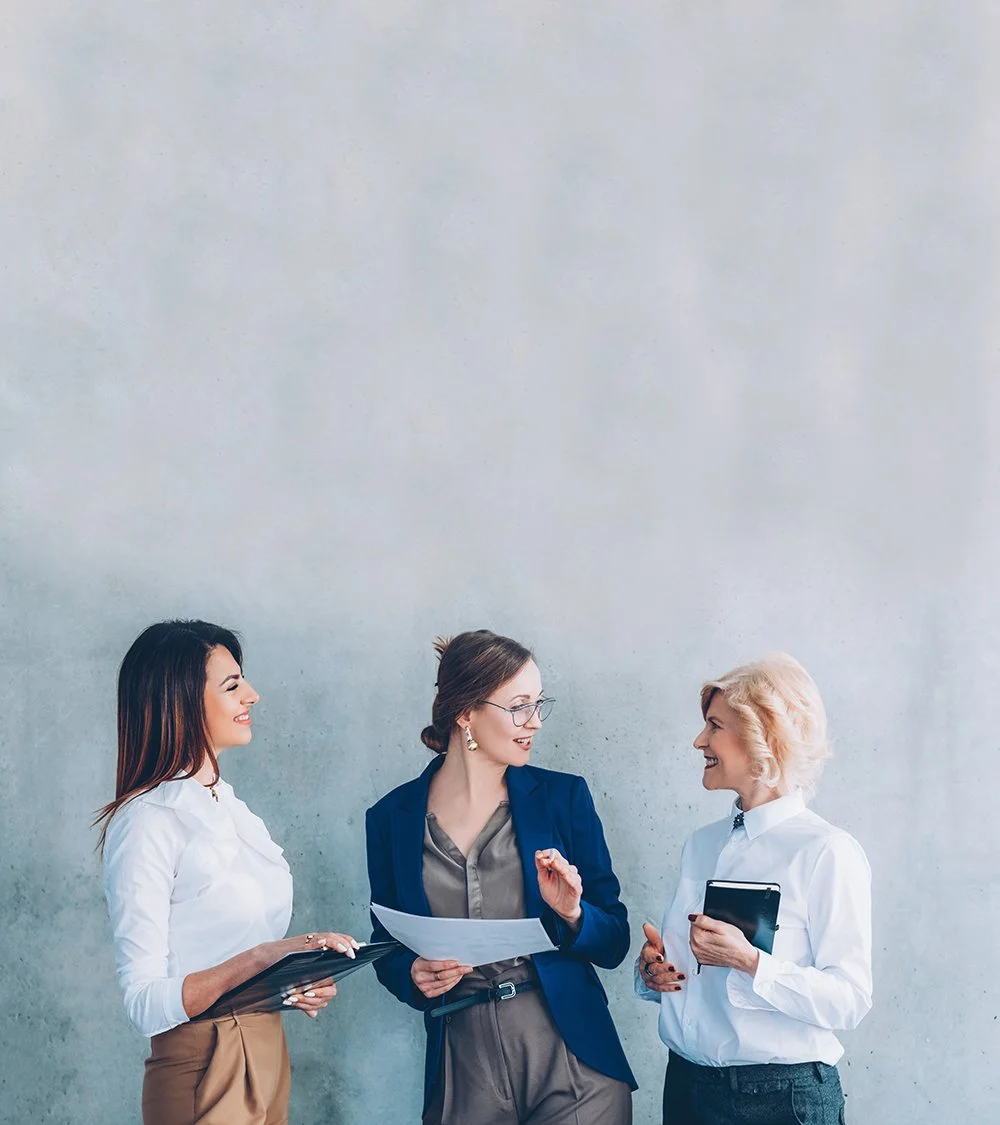Three women in business attire engaged in a conversation against a plain wall.