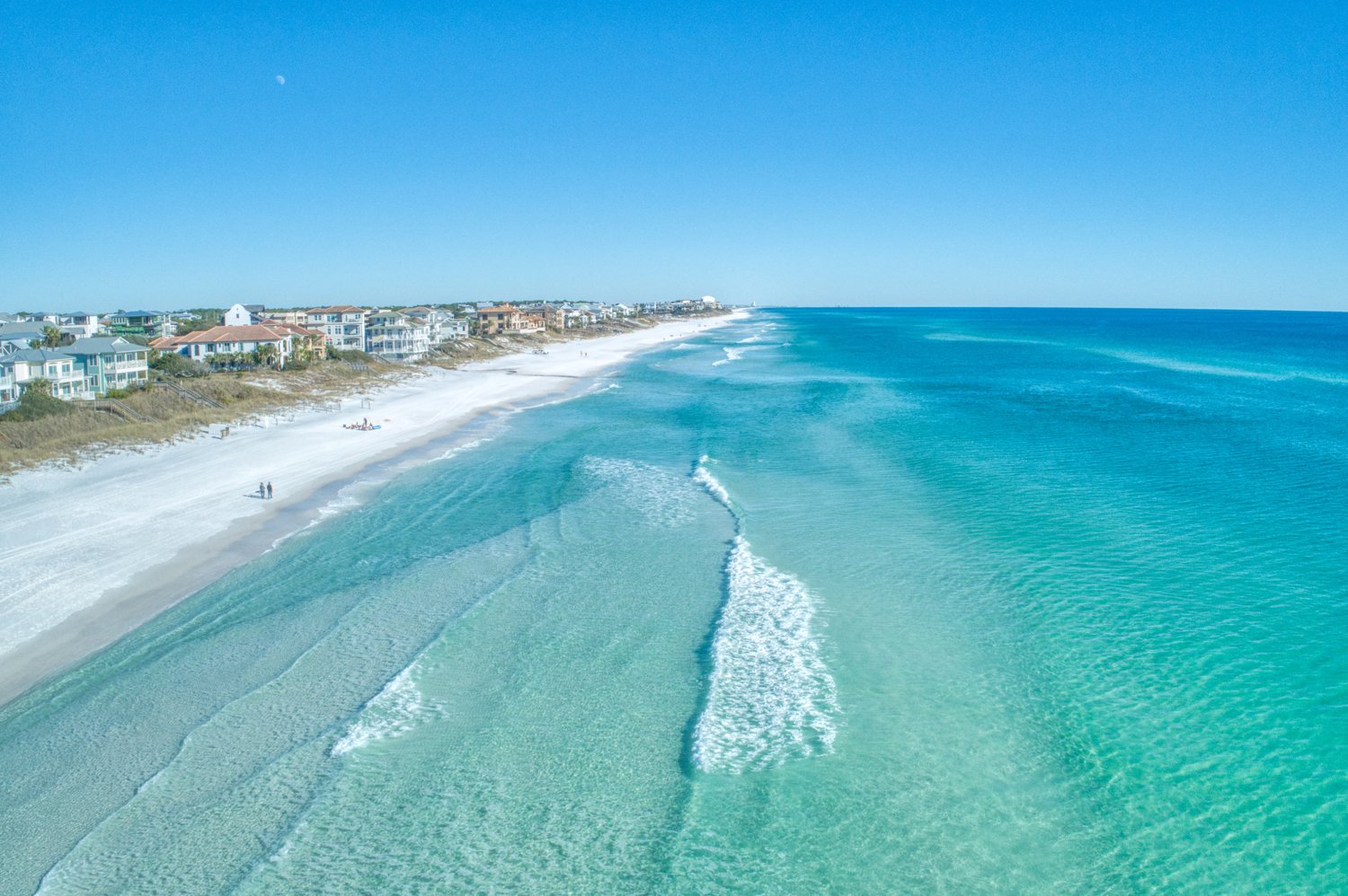 Aerial view of a beach with white sand, coastal houses along the shoreline, and turquoise ocean waves under a clear blue sky.