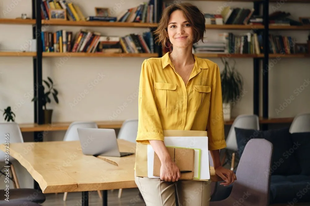 A young woman with short wavy brown hair, wearing a yellow blouse, smiling while holding notebooks and papers in a modern office or library setting, with shelves of books and seating.