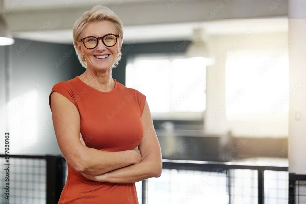 Smiling older woman with short blonde hair and glasses, wearing an orange dress, standing indoors with arms crossed.