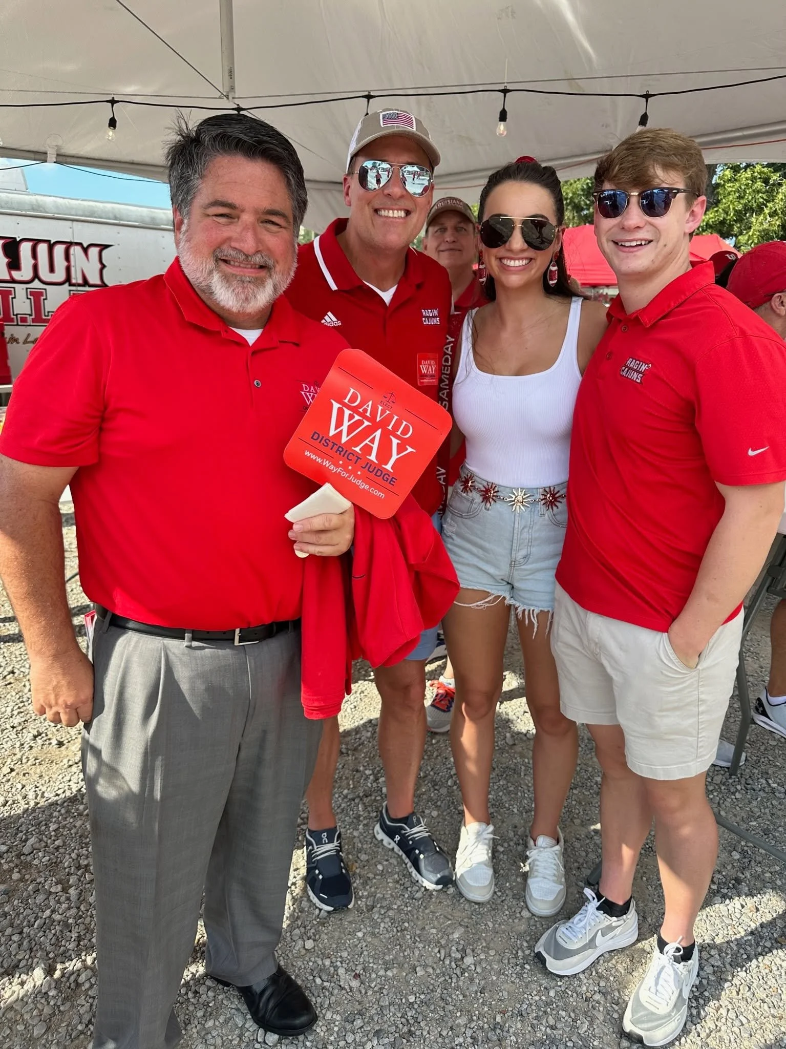 Group of five people standing together outdoors at a daytime event, smiling, with some wearing sunglasses and red shirts, and one woman wearing a white tank top and denim shorts, under a white canopy with string lights.