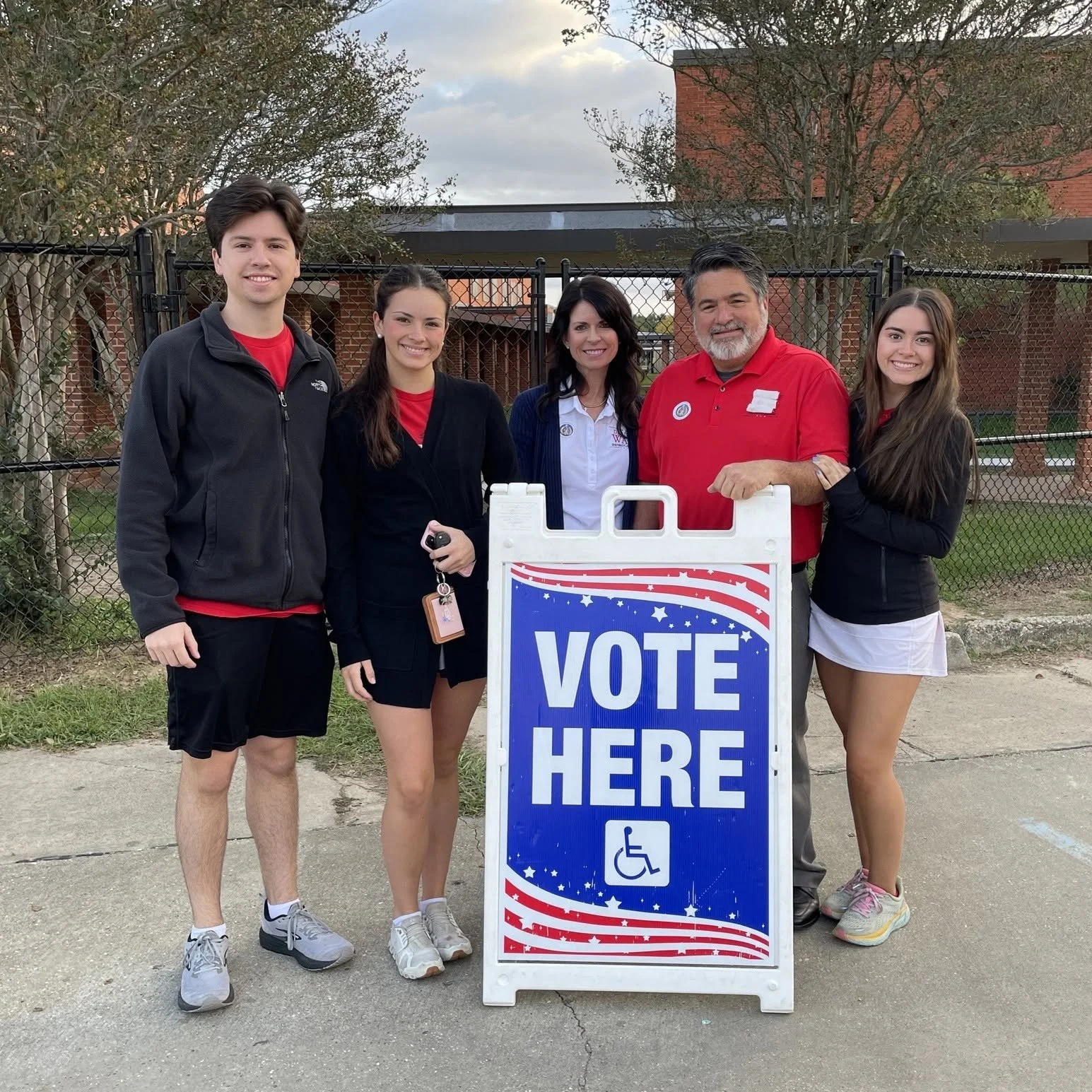 Group of five young people and an older man standing outdoors behind a sign that says 'VOTE HERE' with a wheelchair accessibility symbol. They are smiling and dressed casually, with some wearing red shirts or jackets, in front of trees and a brick bu