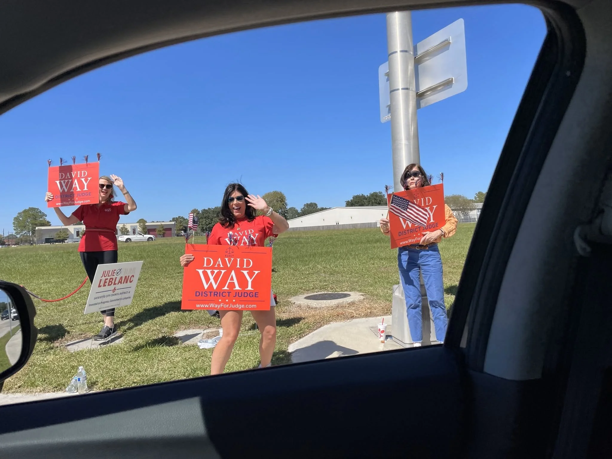 Three women standing on a grassy area holding campaign signs, with one waving and smiling, and two holding American flags, seen through a car window with a blue sky and buildings in the background.