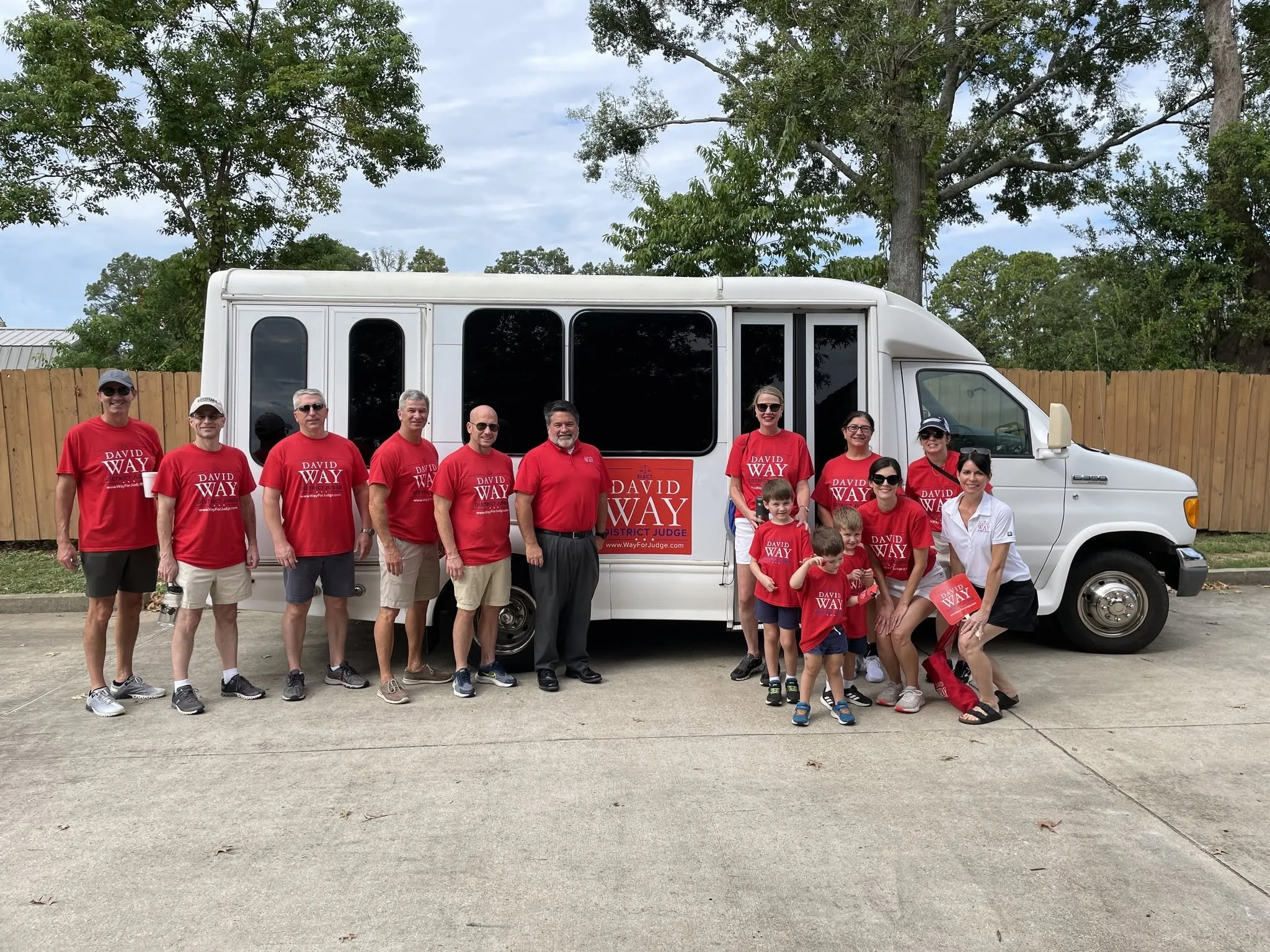 A group of people standing in front of a white vehicle with large windows, some of them wearing matching red shirts with 'David Way' printed on them. The background includes tall trees and a wooden fence.