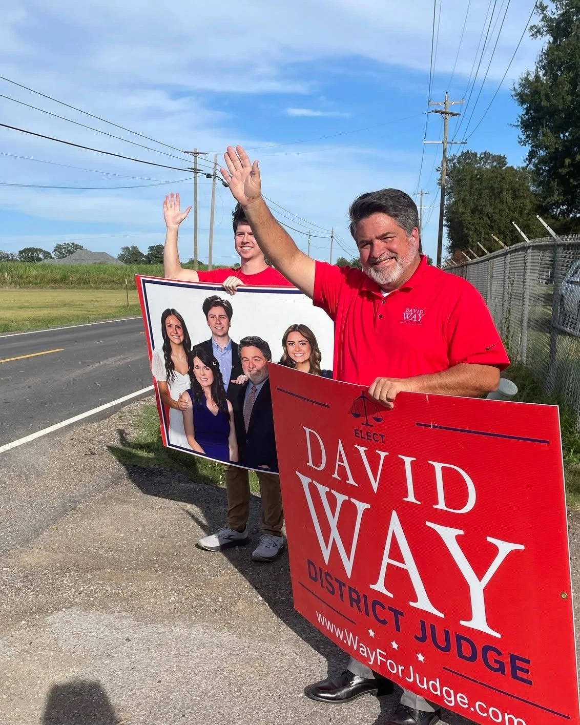 A man and a woman holding campaign signs standing by the side of a rural road with power lines and trees in the background. The man is wearing a red shirt with a campaign logo, smiling, and waving. The signs promote candidate David Way for district j