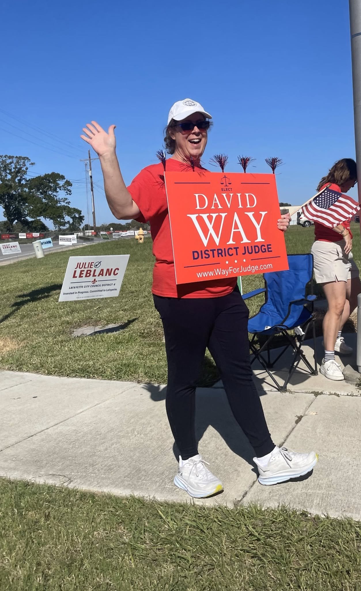 A woman smiling and waving at an outdoor event, holding a sign for a political candidate named David Way, with blue skies and other people in the background.