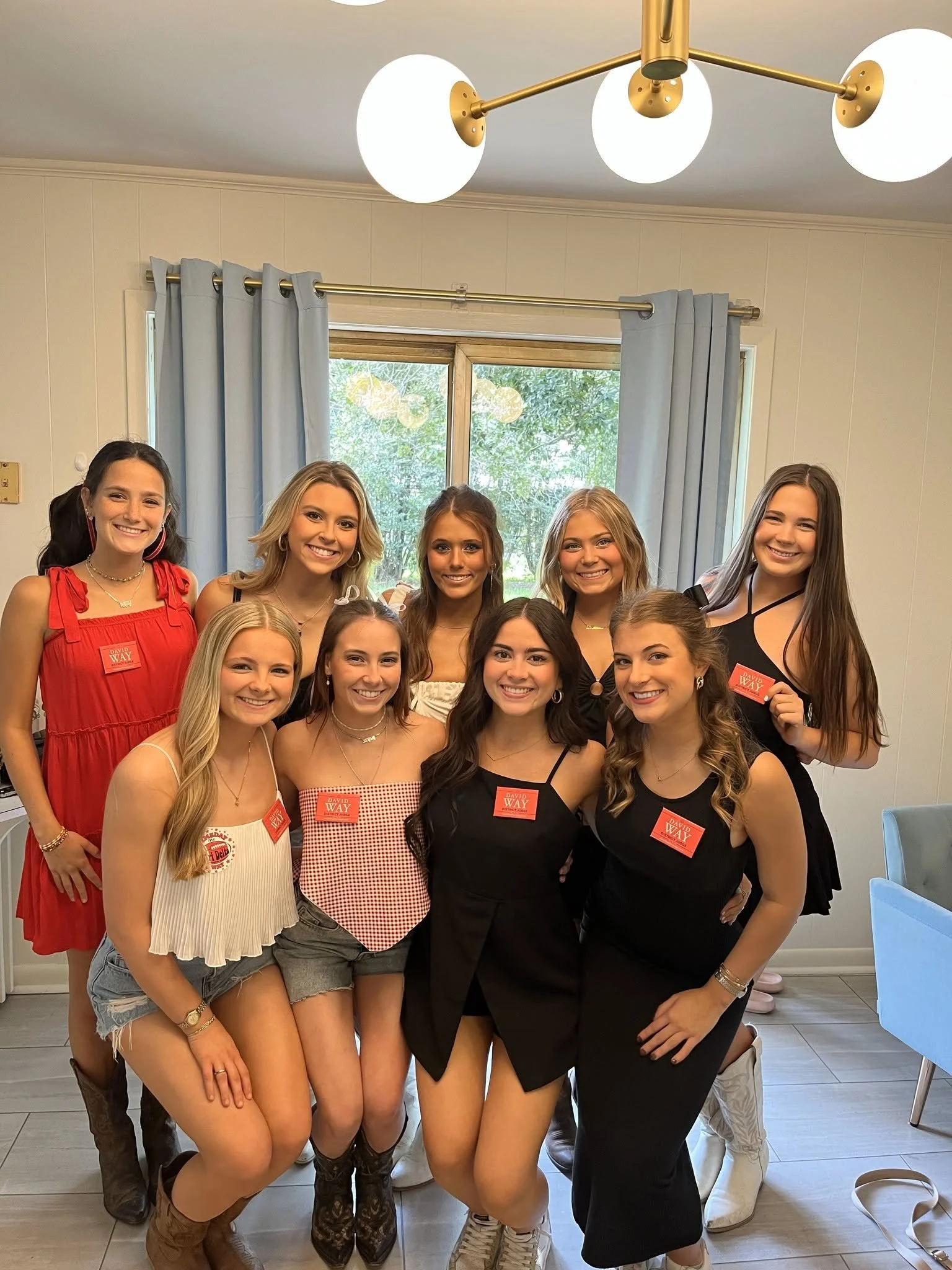 A group of young women standing together inside a house, smiling for the photo. They are wearing casual summer outfits with some wearing name tags, and a window with blue curtains and outdoor greenery is visible in the background.