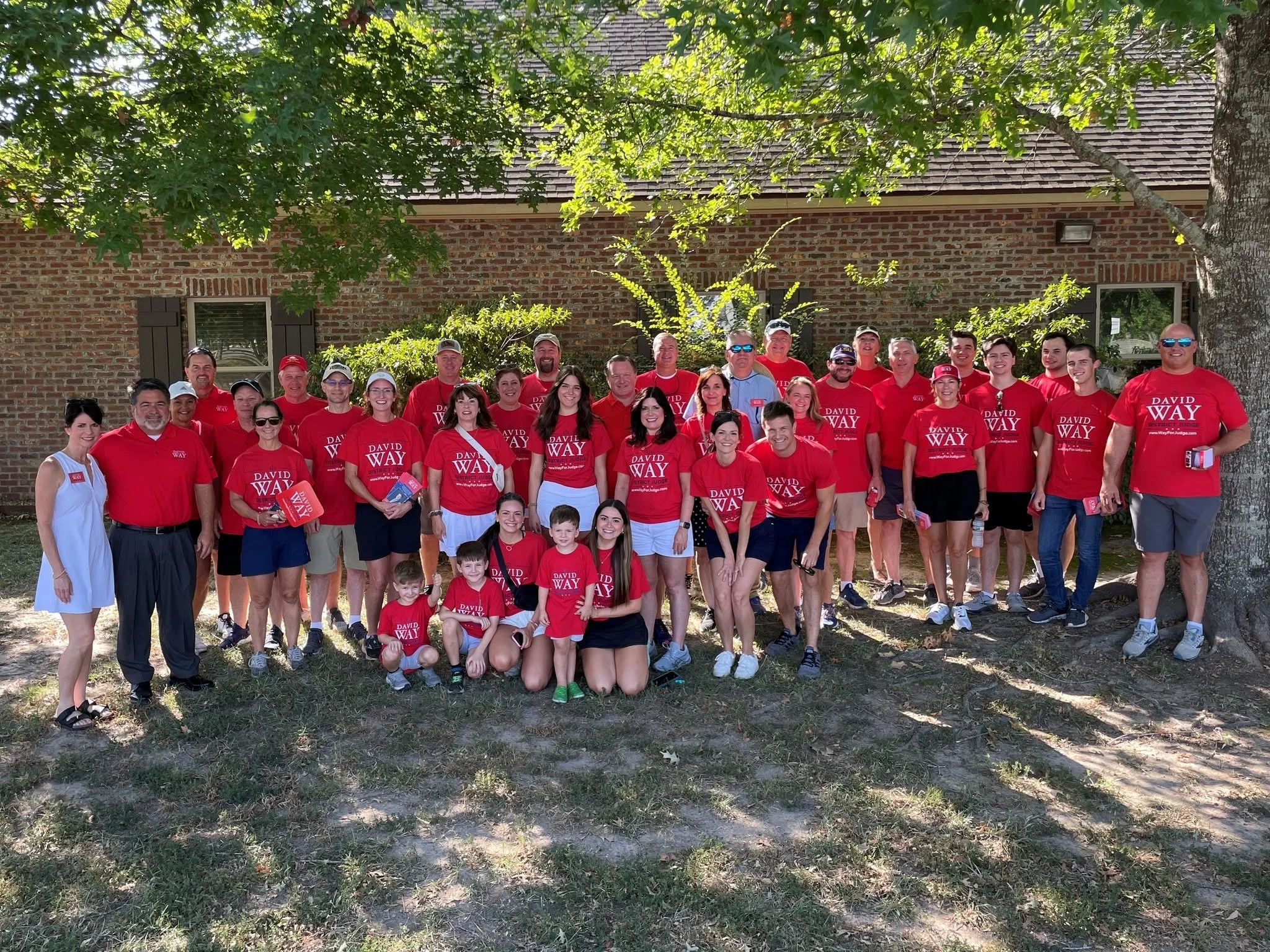 Group of men, women, and children standing outdoors in front of a brick building with a roof and trees, many wearing red shirts with white text that reads 'David Way District Judge,' smiling and posing for the photo.