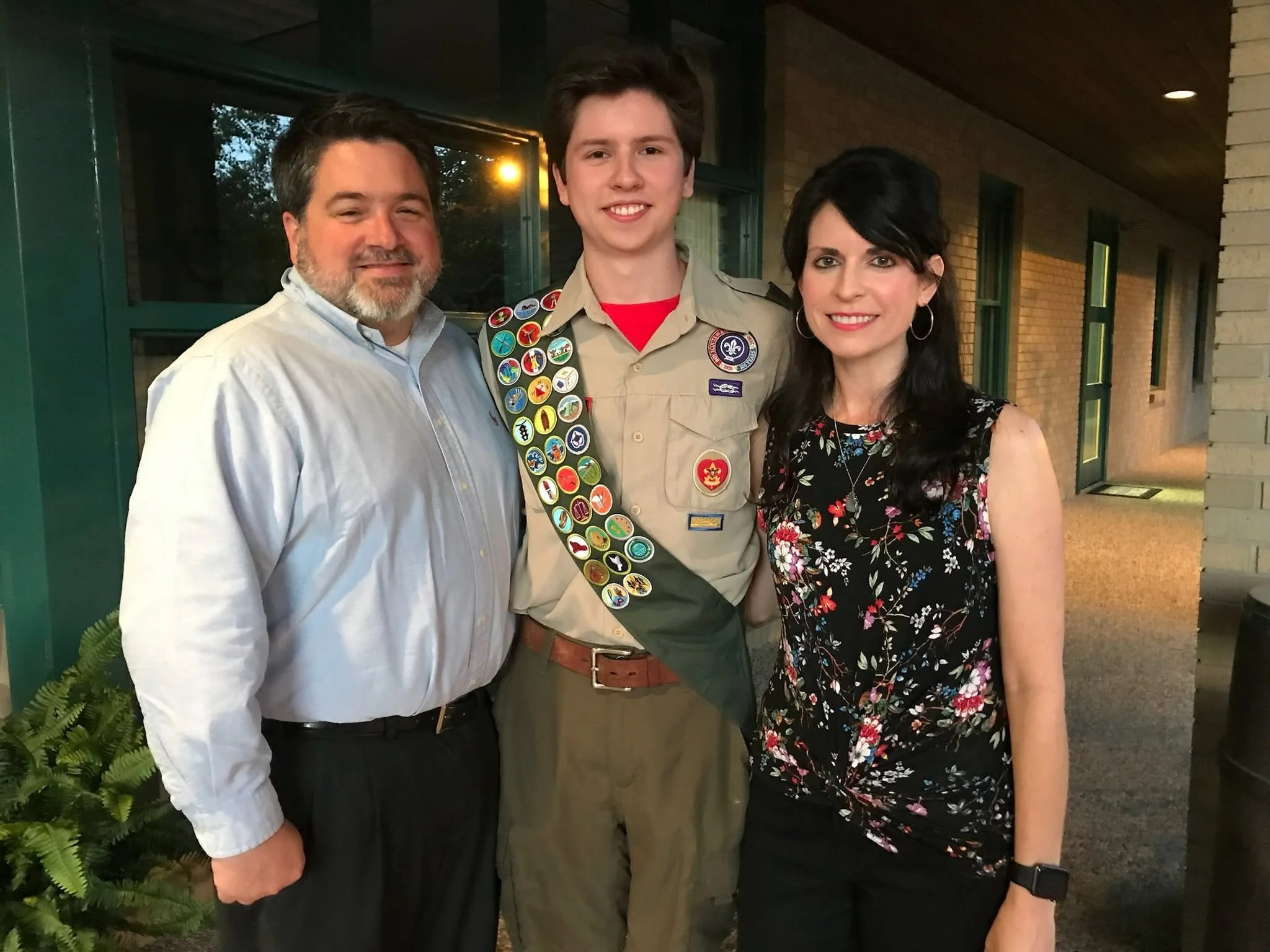 Three people standing close together outside at sunset, smiling. The person in the middle is a young man with wearing a Boy Scout uniform with patches and badges, including a sash decorated with various badges. The person on the left is an older man 