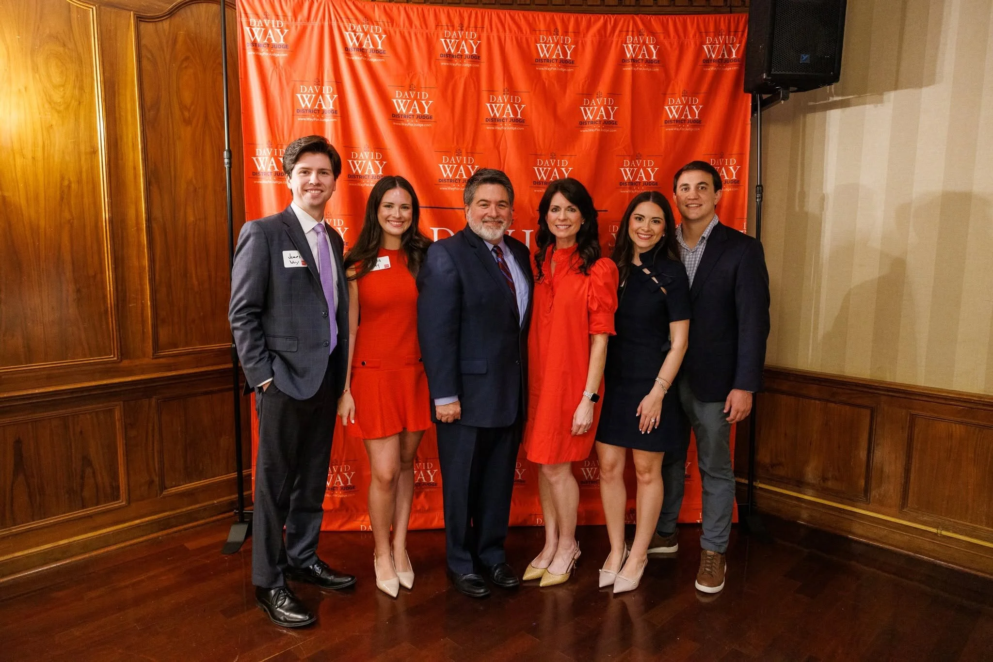 Group of six people standing together in front of a red backdrop with the name 'David Way District Judge' repeated on it, dressed in formal attire, smiling for the camera in a wood-paneled room.