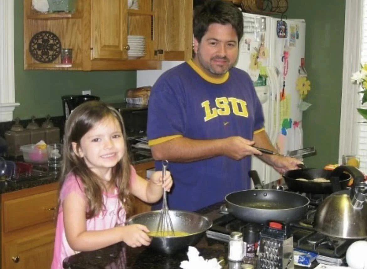 A man with dark hair and a beard wearing a blue LSU t-shirt is cooking on a stove. A young girl with long brown hair wearing a pink striped shirt is whisking eggs in a bowl, smiling at the camera.