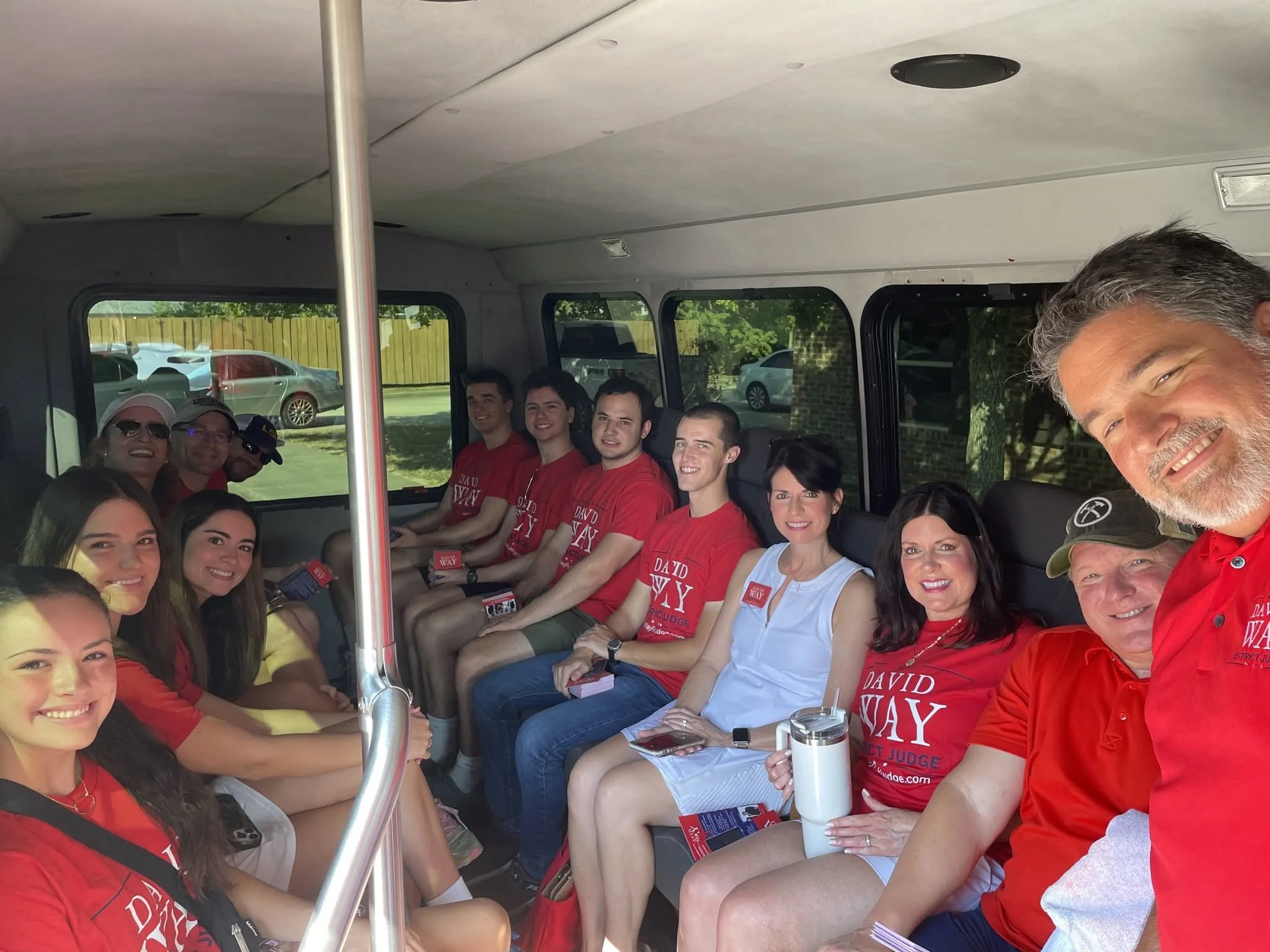 A group of diverse people sitting inside a bus or van, smiling and wearing red shirts. They are part of a group event or celebration. Outside the window, cars are parked.