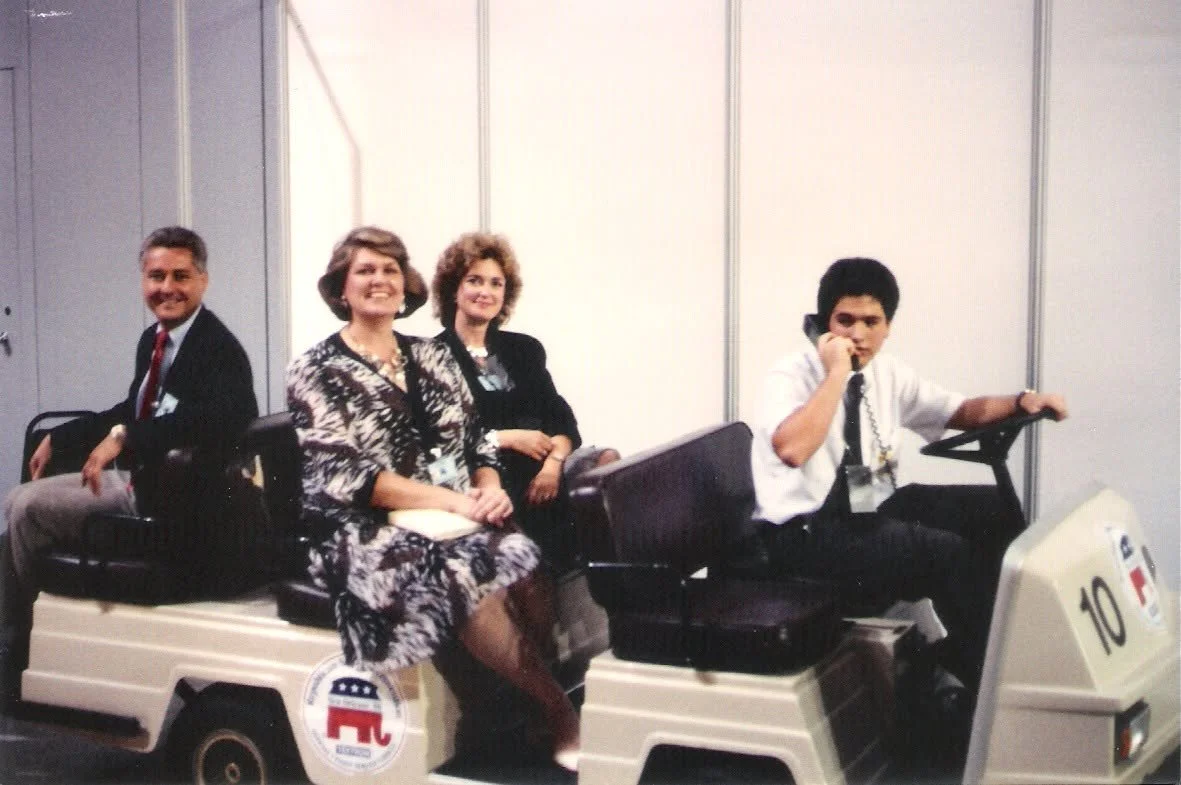 Four people sitting on a small cart with an 'R' logo, with three women and one man on a phone, in an indoor setting.