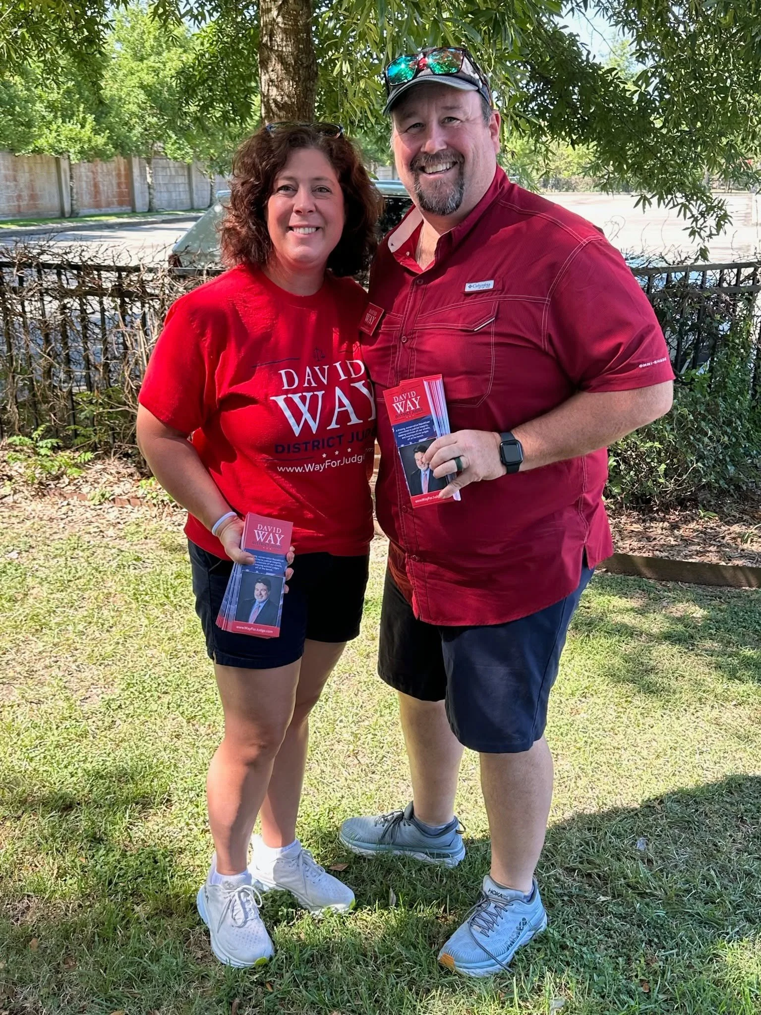 A smiling woman wearing a red campaign t-shirt and a man in a red shirt holding campaign flyers. They are outside under a tree on a grassy area with a fence and parked car in the background.