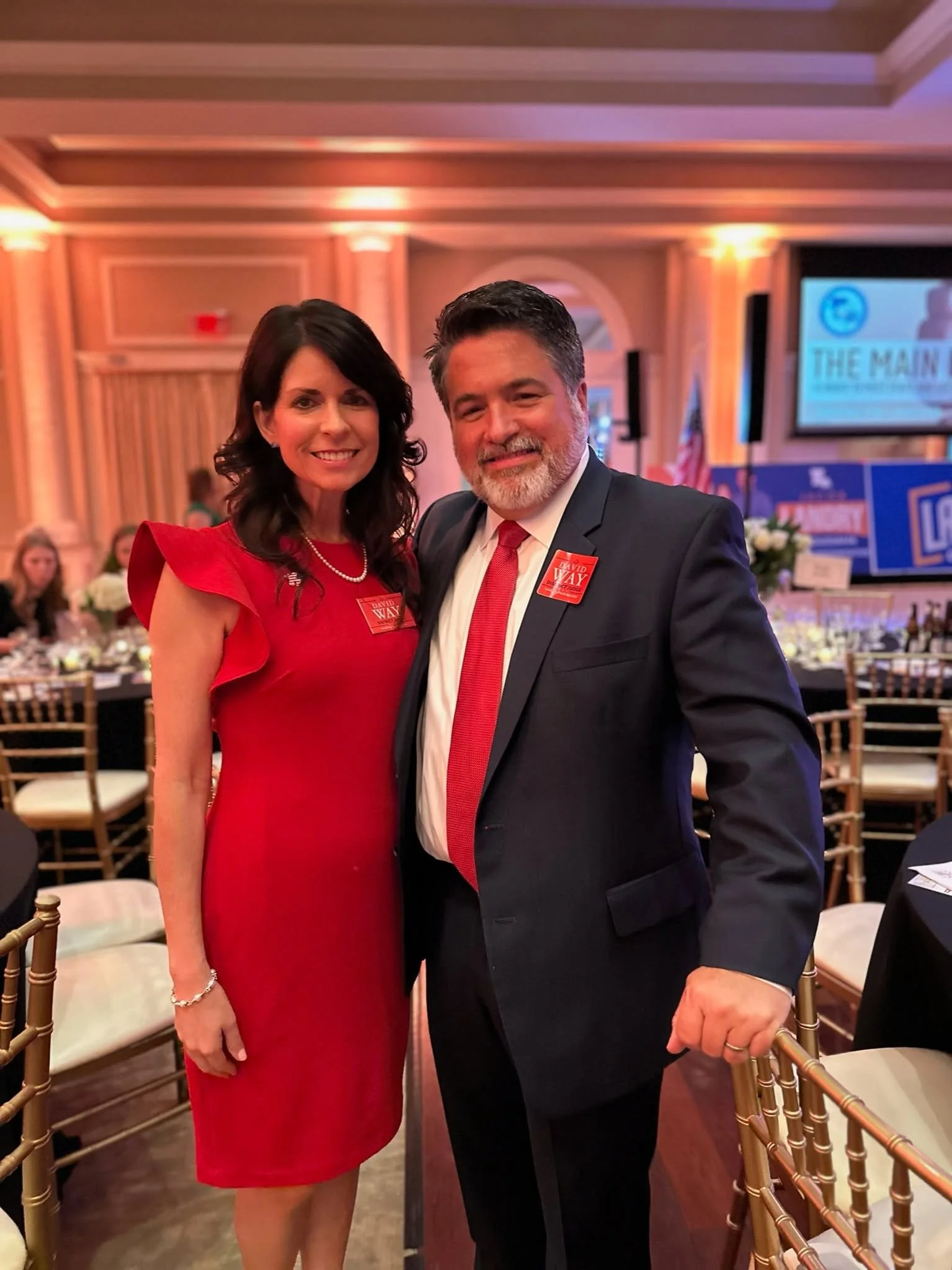 A woman in a red dress and a man in a black suit with a red tie smiling and posing together at a formal event with a decorated ballroom in the background.
