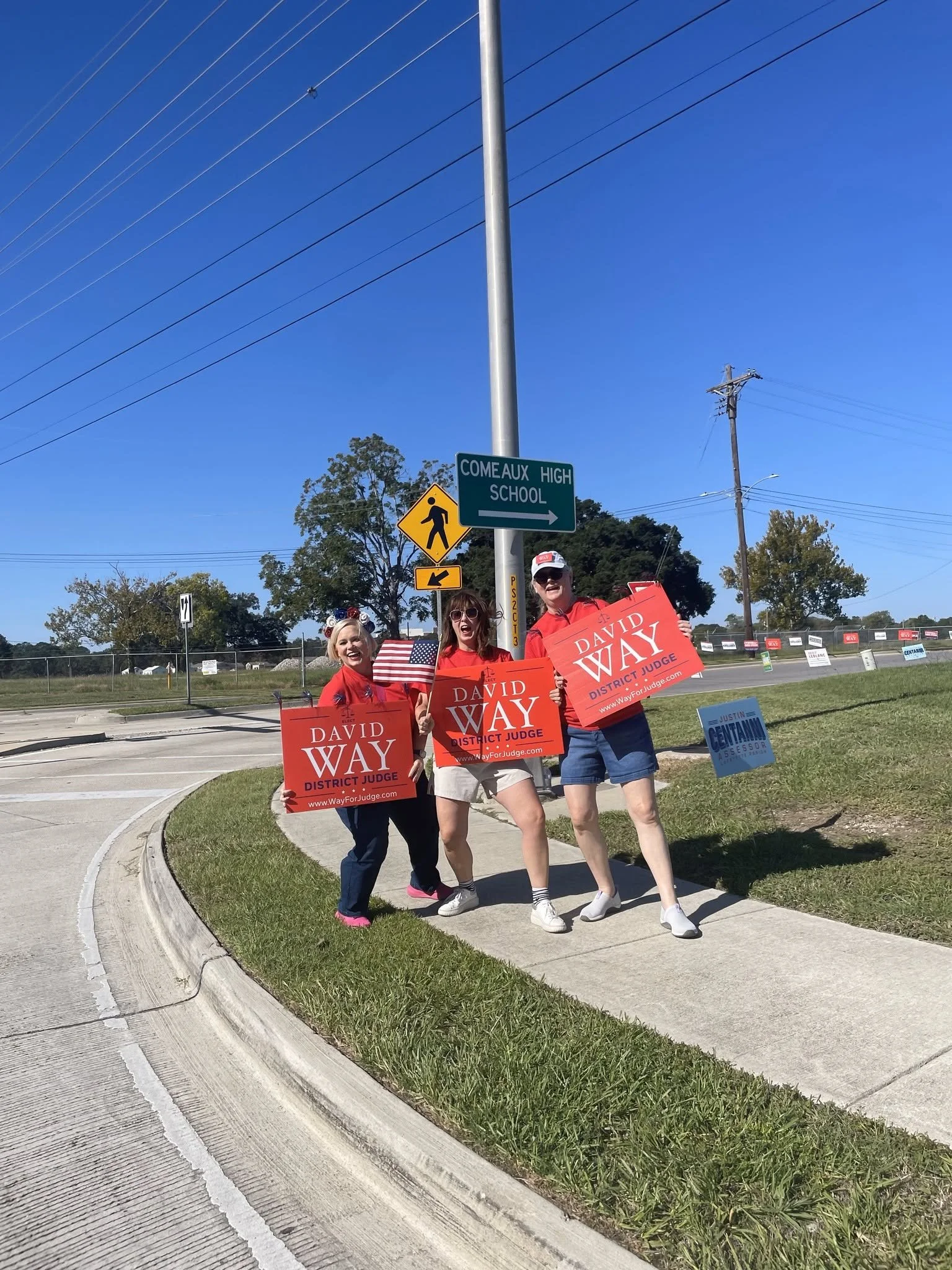 Three women holding campaign signs and waving flags on a sidewalk near a street with signage for Comeaux High School and political campaign signs in the background.