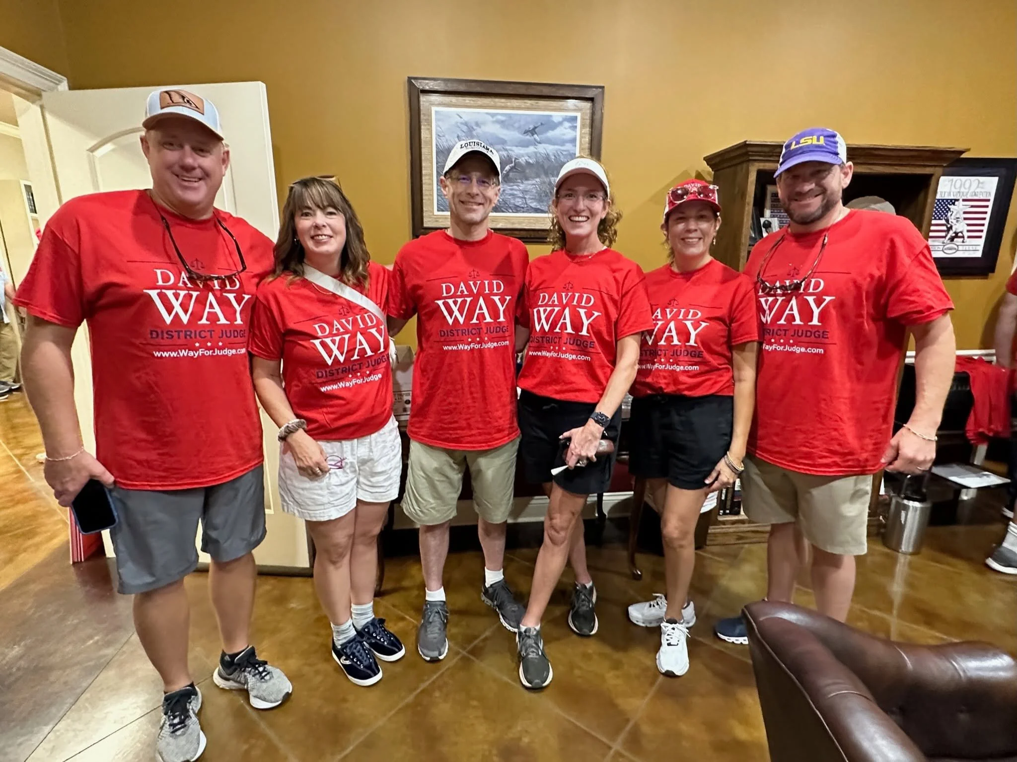 Group of six people wearing red campaign T-shirts with 'David W. Way, District Judge' printed on them, standing inside a room with yellow walls, wooden furniture, and framed pictures, smiling at the camera.