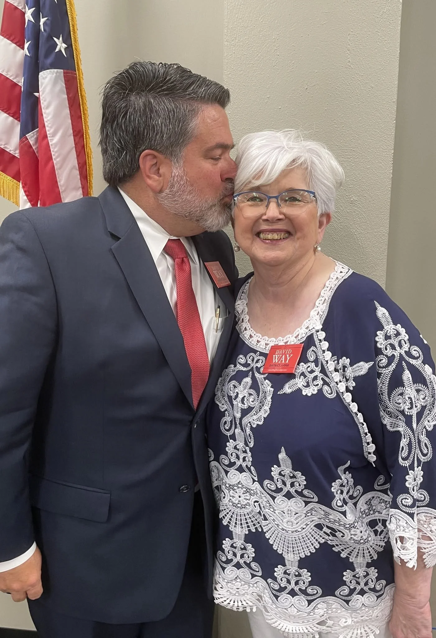 A man in a dark suit and red tie kisses a woman with white hair and glasses on the cheek. The woman is wearing a navy blue dress with white lace embroidery and has a red name tag. An American flag is partially visible in the background.