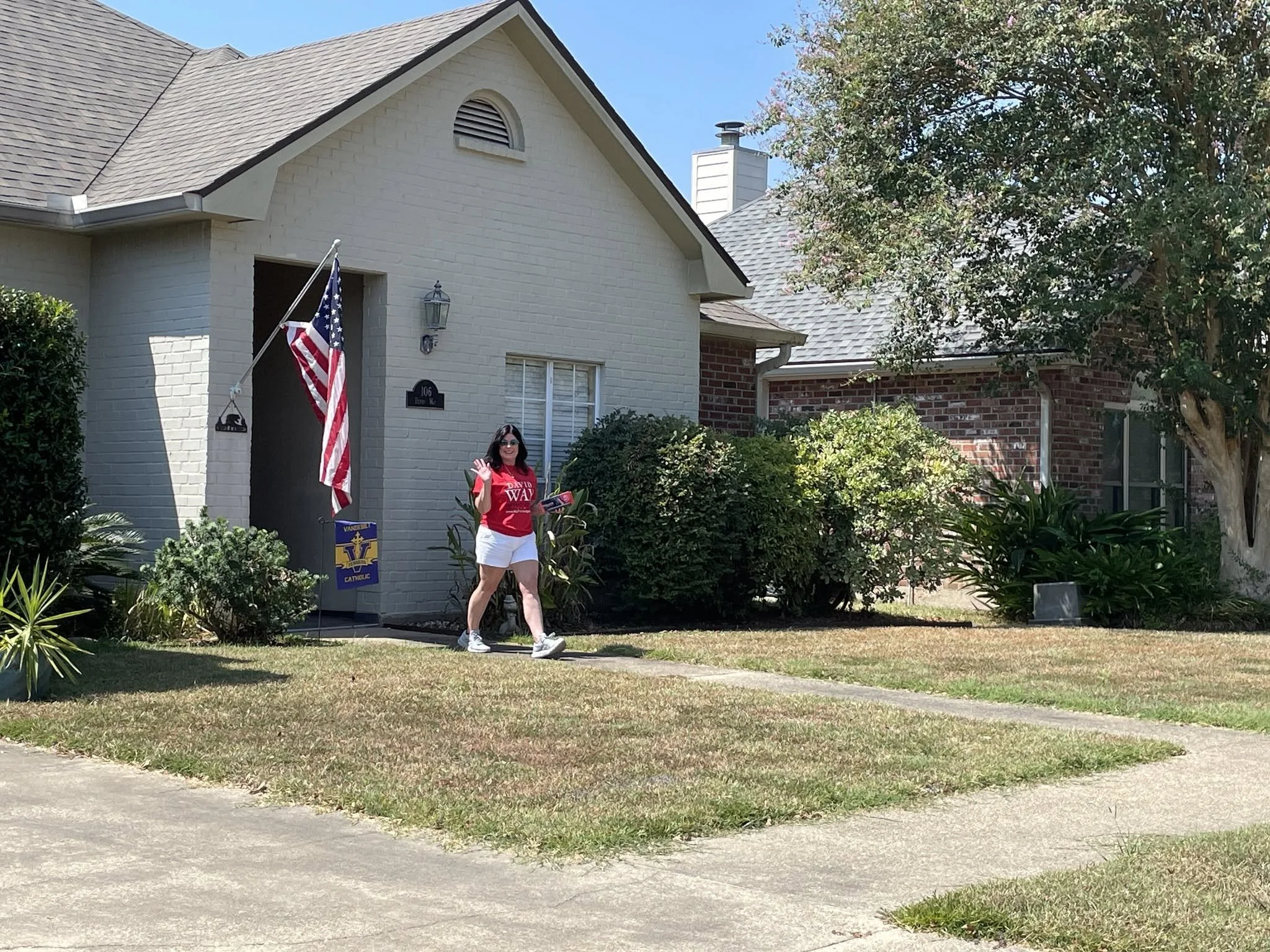 A woman wearing sunglasses and a red campaign t-shirt waving and holding a pamphlet in front of a house with American flags on the porch.