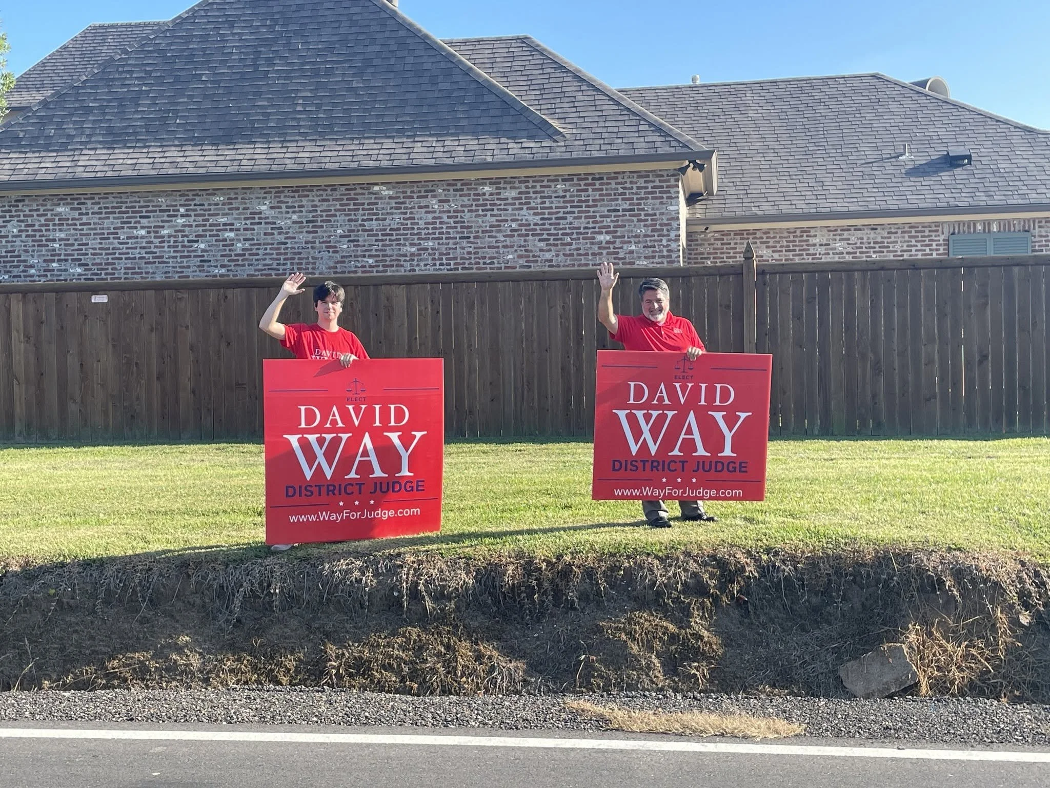 Two people standing behind yard signs that read 'Elect David Wray District Judge' in front of a brick house with a wooden fence, clear blue sky, and green grass.