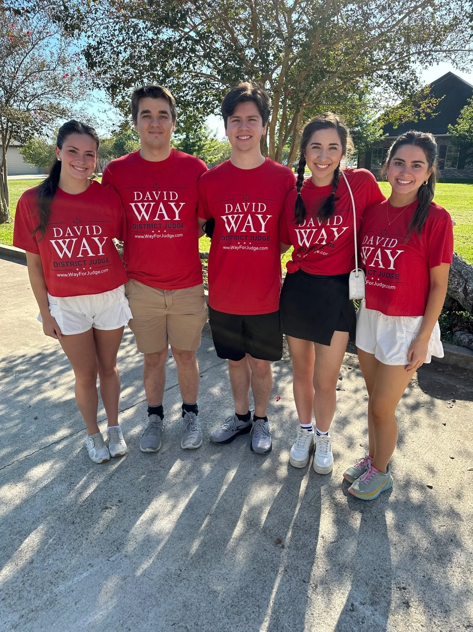 Five young people standing outdoors on a sunny day, all wearing red shirts with white text that reads 'David Wray District Judge' and a web address. They are smiling and standing close together, with trees and a house in the background, casting long 
