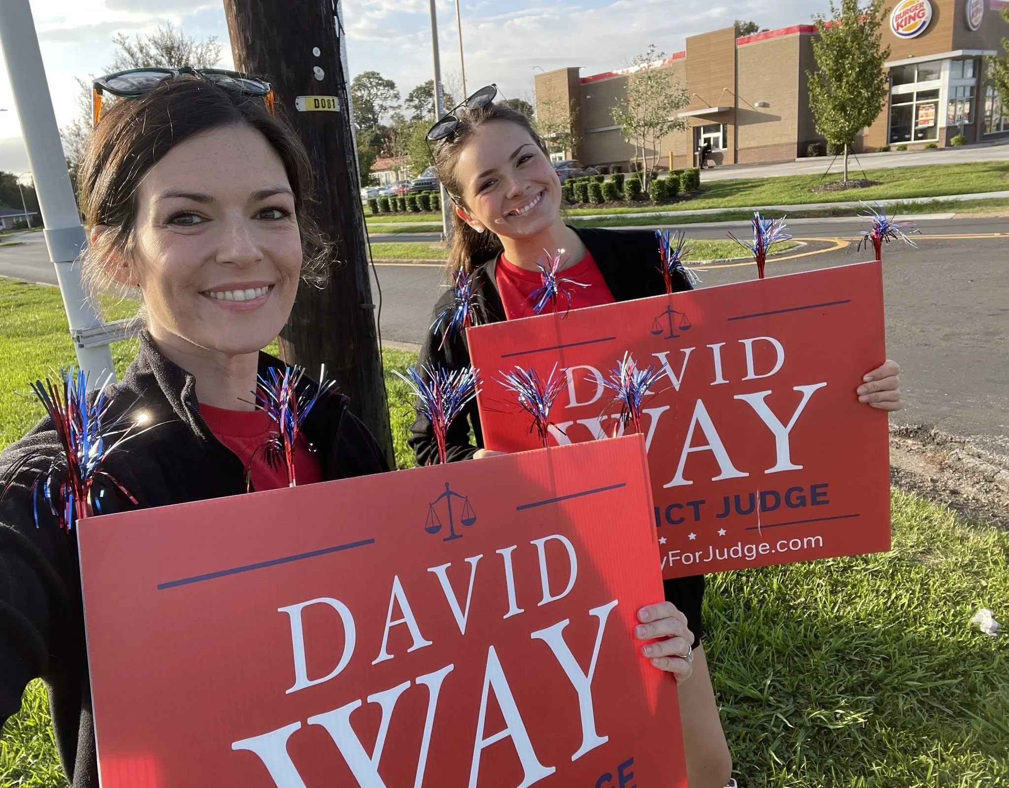 Two women smiling and holding red signs that say "Did Not VOTE" outside near a Burger King restaurant. One woman has glasses on her head and wears a black jacket, the other is in a red shirt and has a backpack. They are decorated with red, white, and