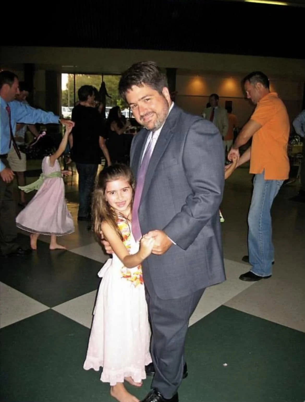 A man in a gray suit and a young girl in a white dress dancing together at a party or celebration, with other couples dancing in the background.