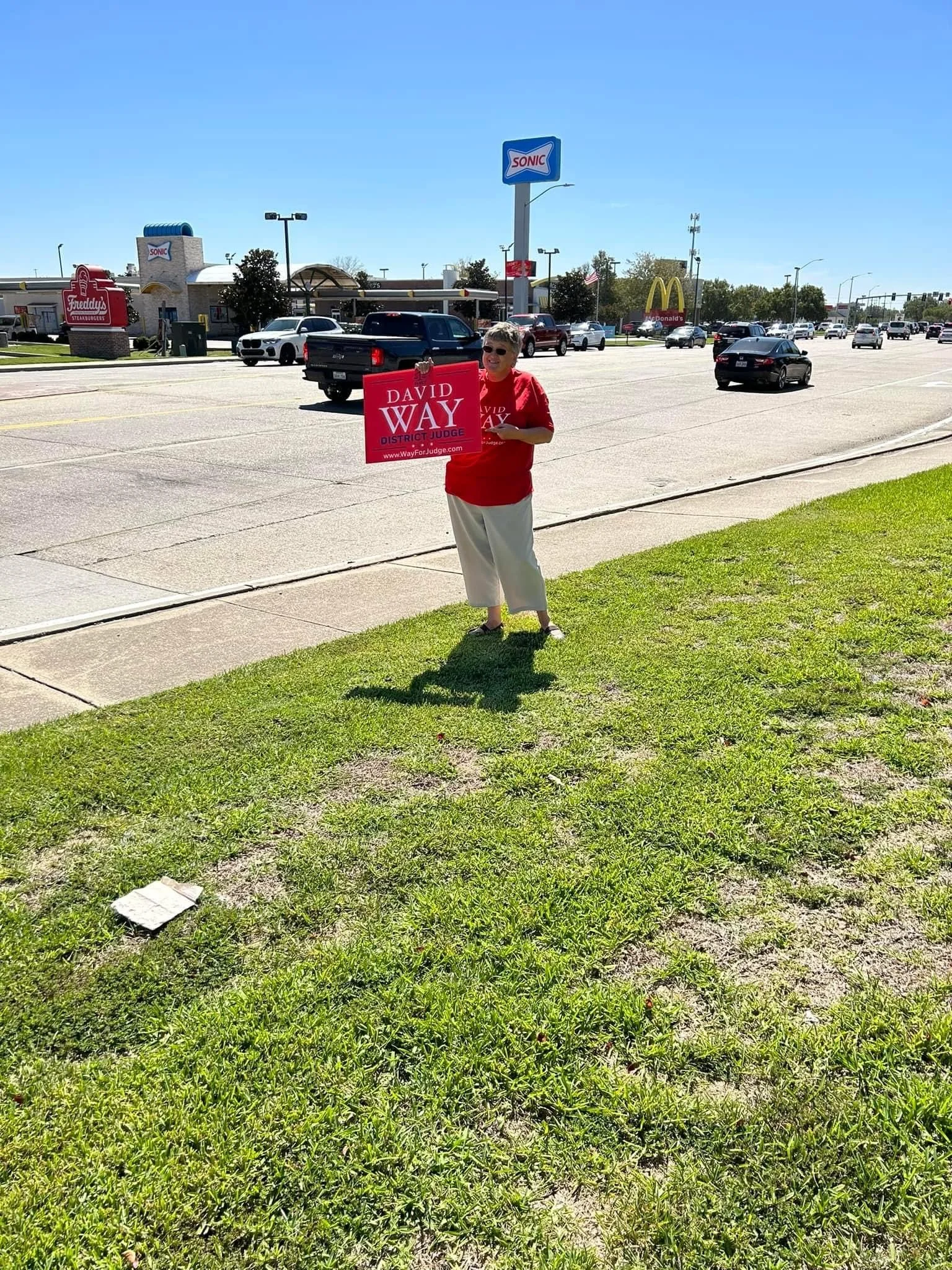 A woman wearing a red shirt and beige pants holding a red campaign sign that says 'David Way District Judge' stands on a grassy area beside a busy street with cars and commercial buildings in the background.