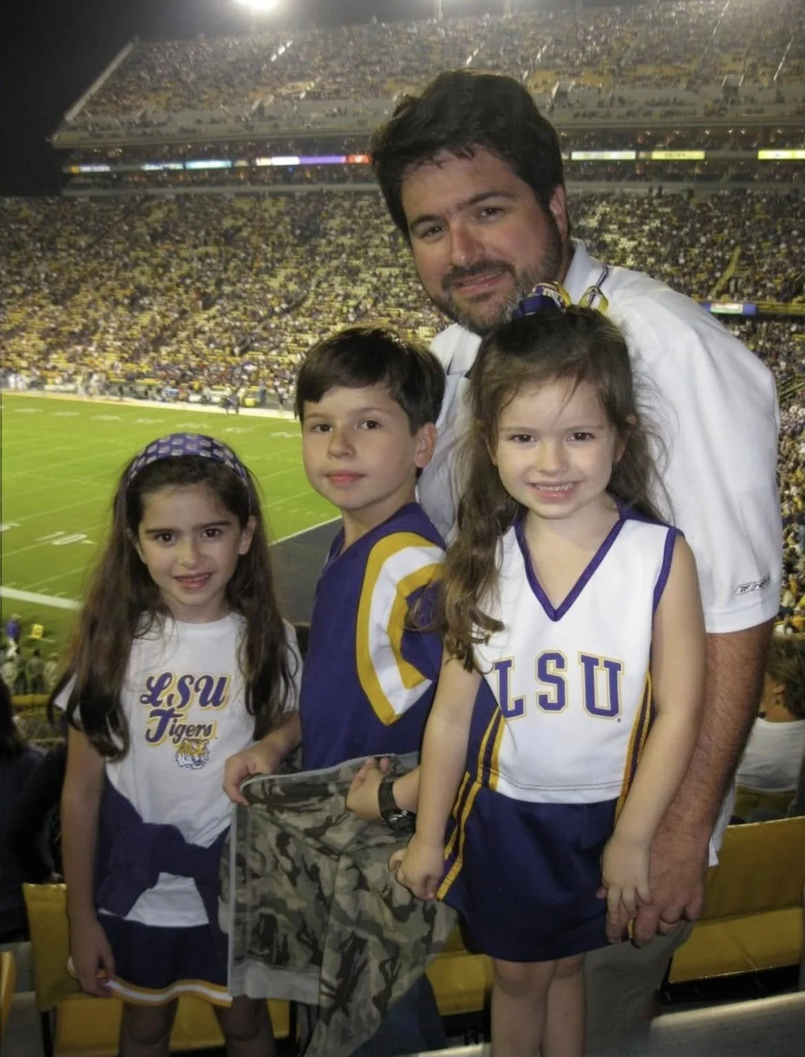 A man and three young children at a football game in a stadium, wearing LSU game jerseys, with the field in the background.