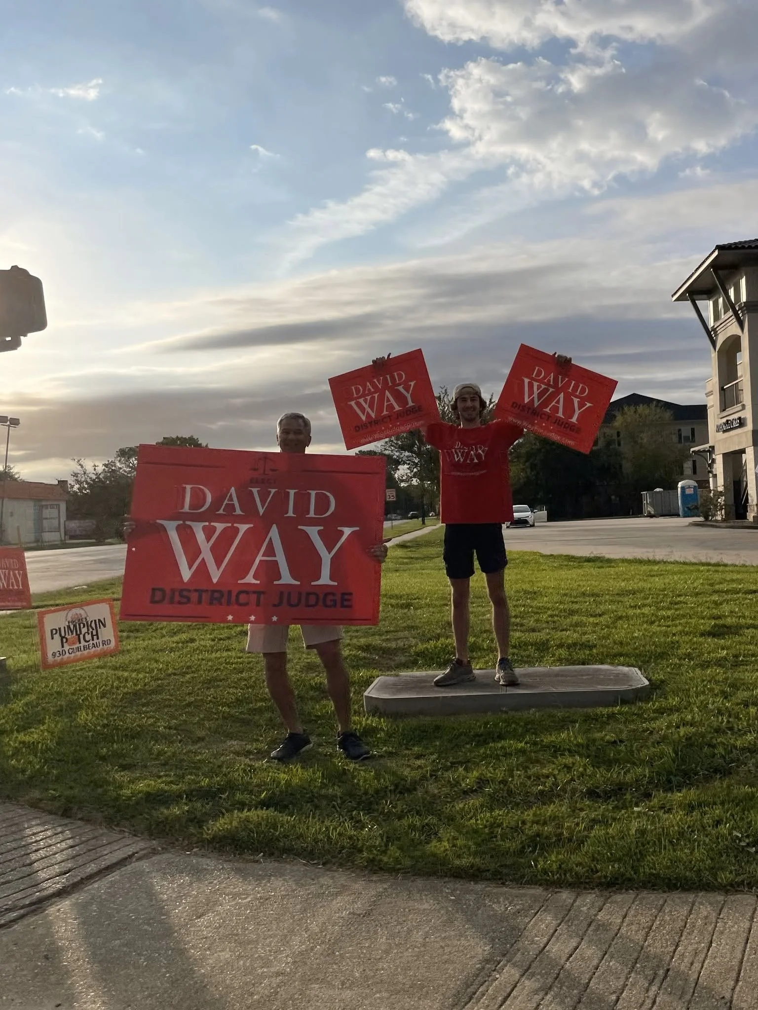 Two men are standing outdoors holding signs that display support for David Way, a candidate for district judge. They are in a grassy area near a sidewalk, with buildings and parked cars visible in the background under a partly cloudy sky.
