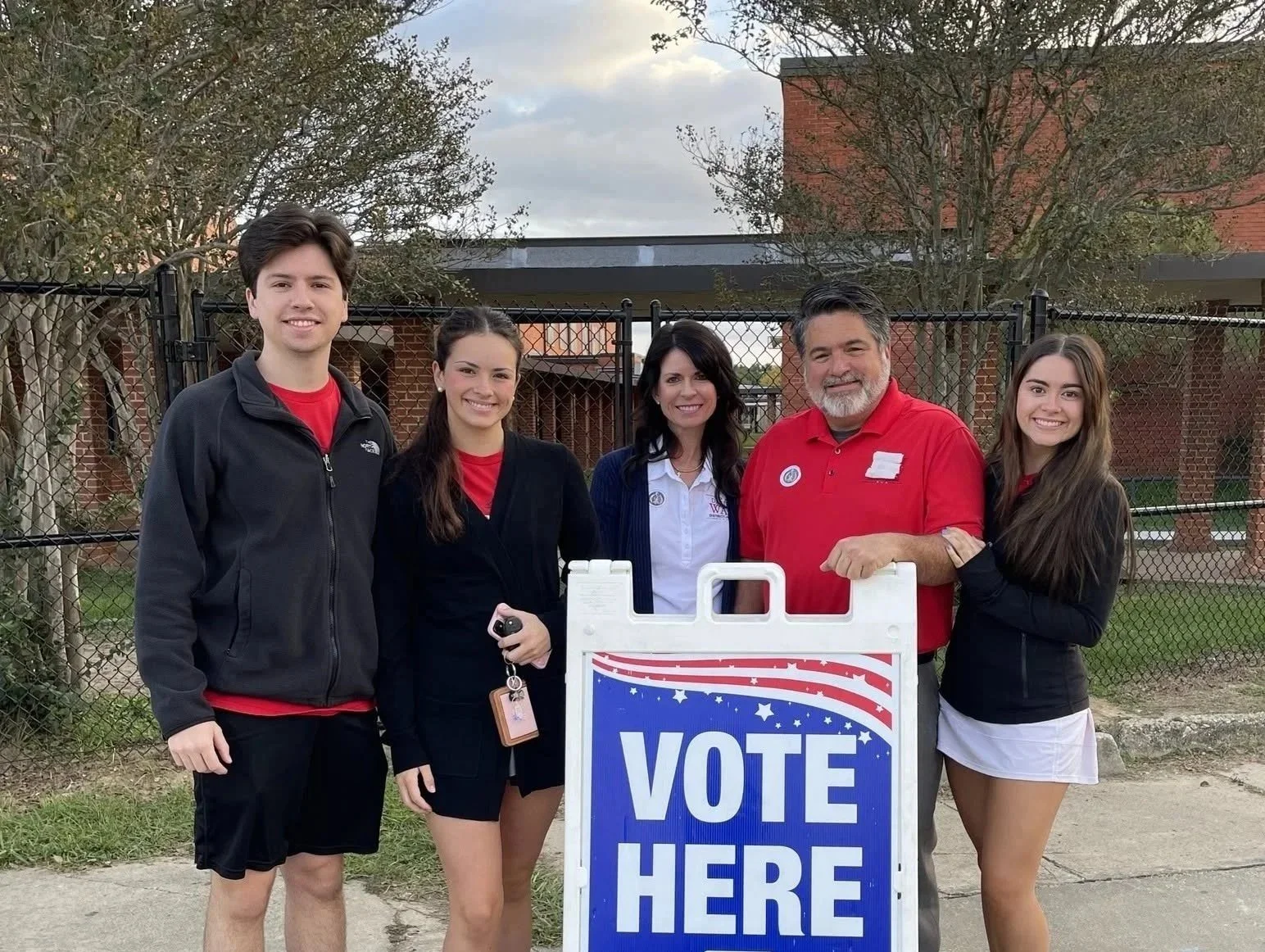 David Way and family standing outside behind a fence, smiling, with a sign that says 'Vote Here.' The group includes three women and two men, dressed in casual and semi-formal attire, participating in a voting or election event.