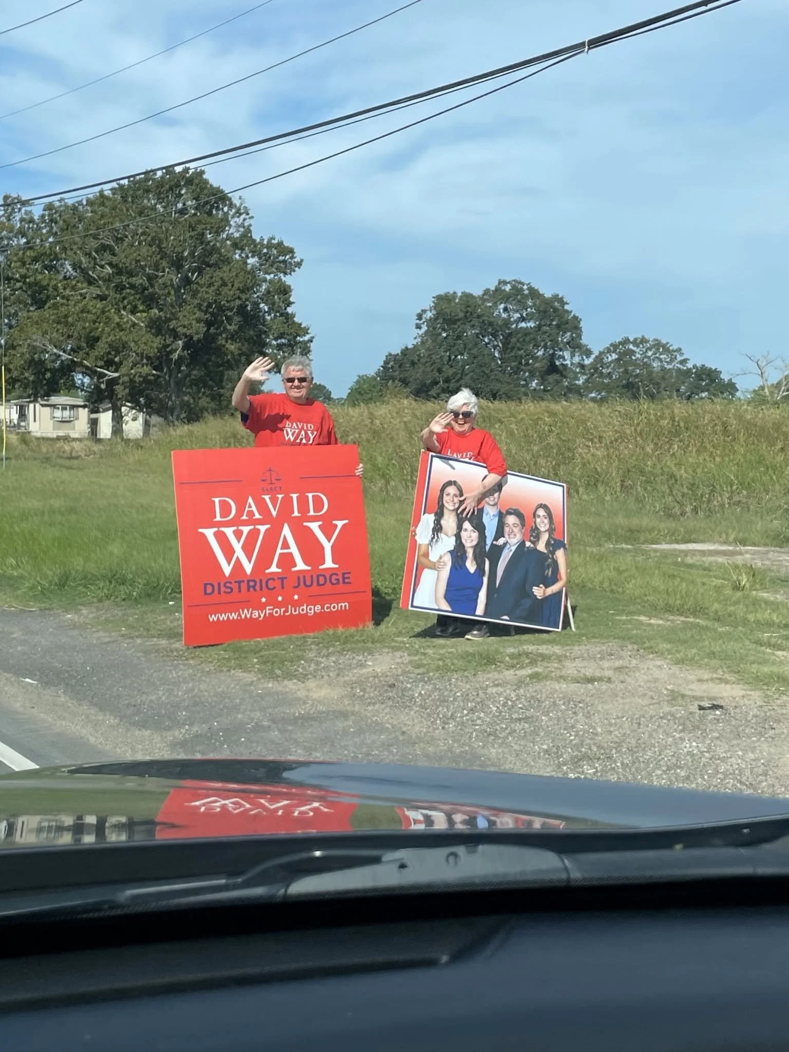 Two people standing on the side of the road at a political rally or campaign event, waving. They are holding signs supporting David Way for district judge. The signs are red and feature campaign images and slogans. The people are wearing red shirts a
