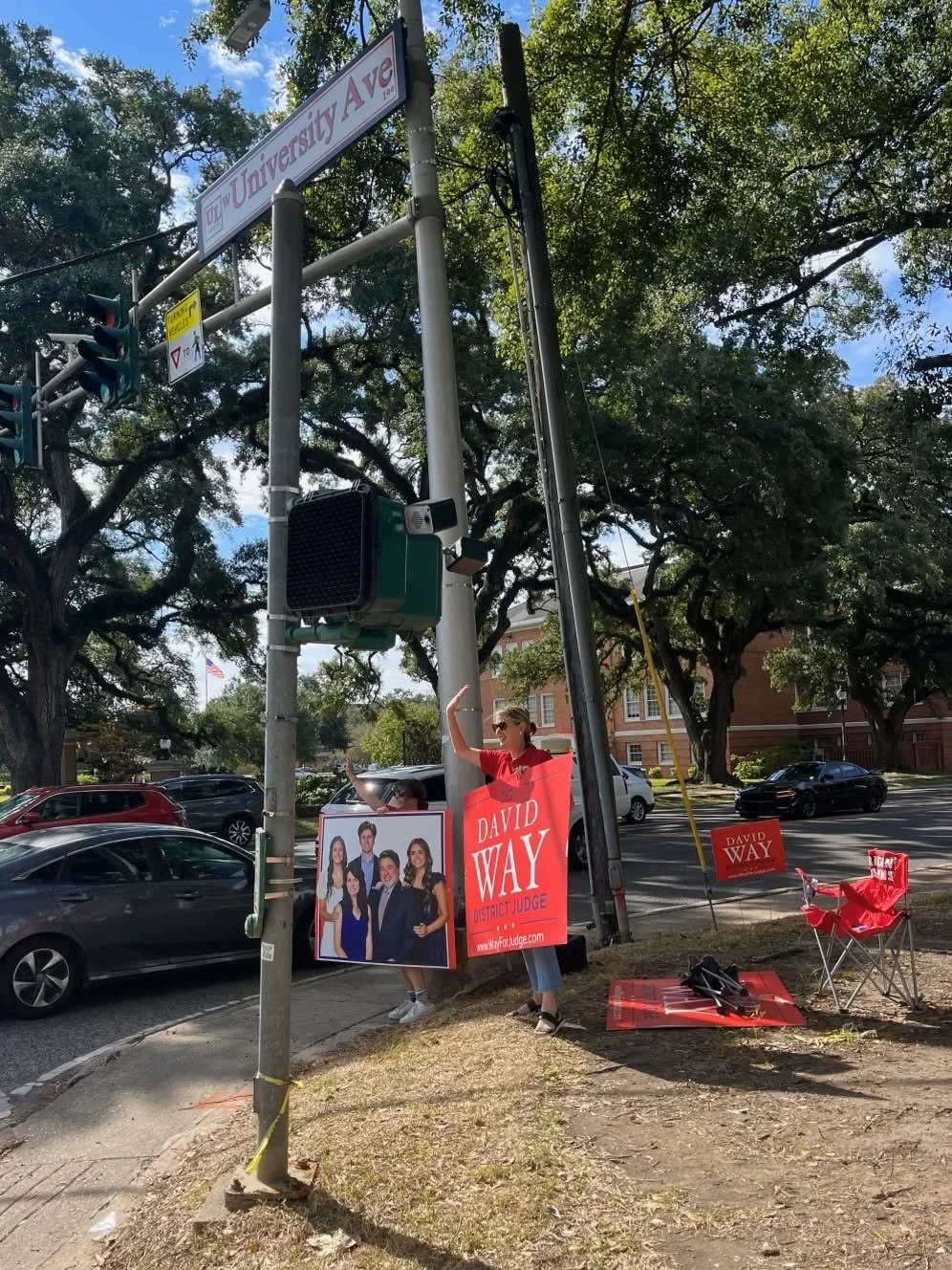 A person standing on a sidewalk holding campaign signs for David Way, a district judge candidate, near a street with cars and trees. The signs display a group photo of candidates and campaign slogans.