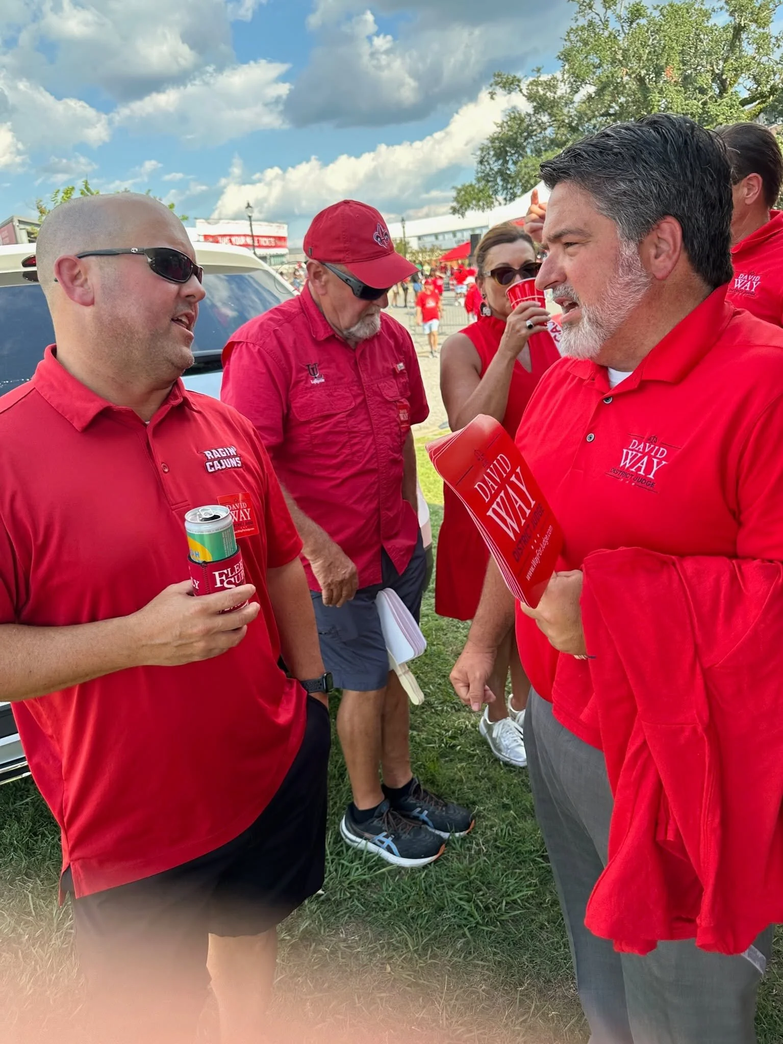 A group of people wearing red shirts, standing outdoors on grass, engaged in conversation during daytime with a cloudy sky and trees in the background. Some individuals are holding red cups and pamphlets, and one person is wearing sunglasses.