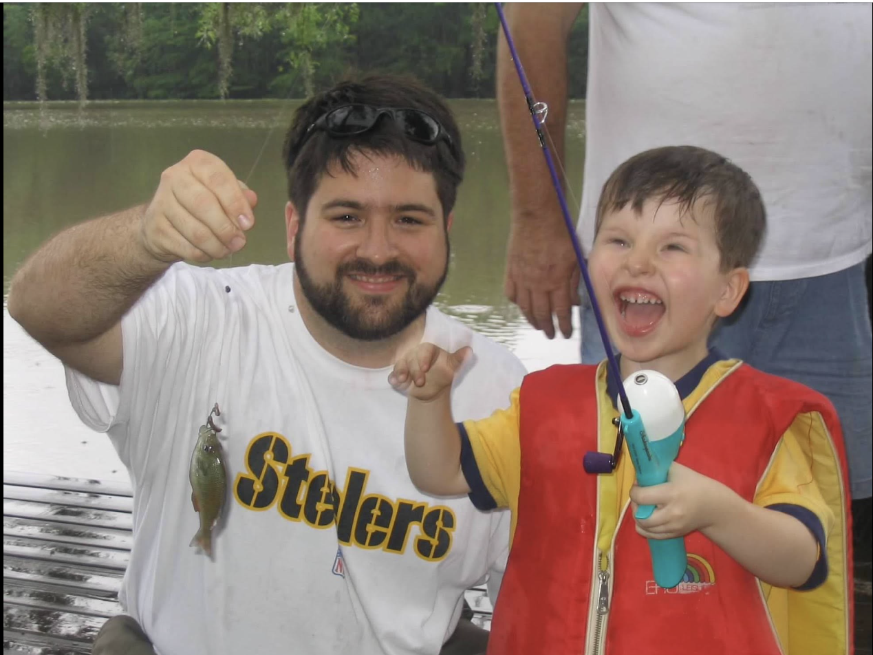 A young boy holding a fishing rod and a fish, smiling, sitting by a lake, with an adult male in the background.