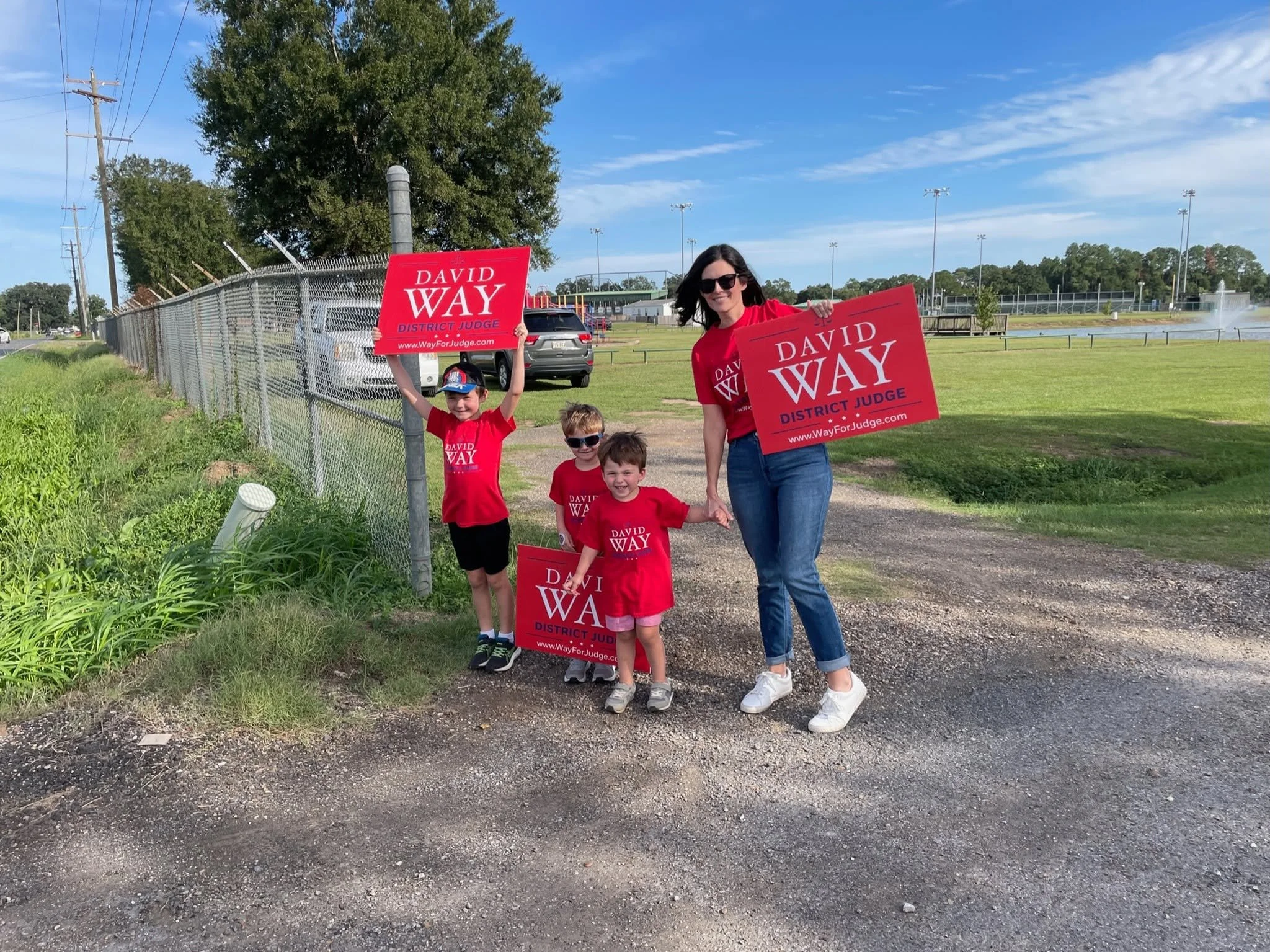 A woman and four children standing outdoors holding campaign signs for David Way, District Judge. The group is near a chain-link fence, with a grassy field, a large tree, and a sports field with lights in the background on a sunny day.