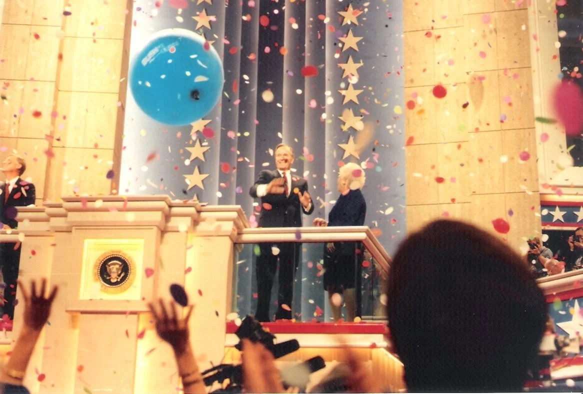 People celebrating at an event with confetti, a large blue balloon, and patriotic decorations including stars and an official seal on the podium